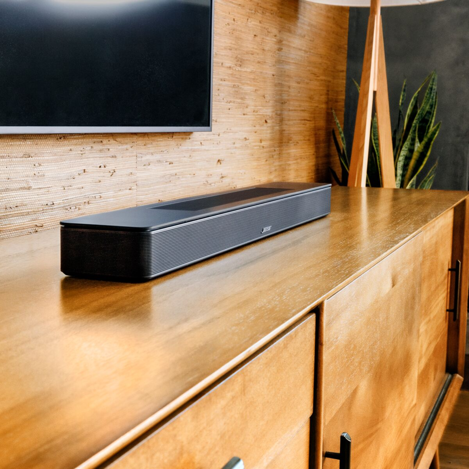 A black soundbar on a wooden cabinet in a room with a TV and plant.