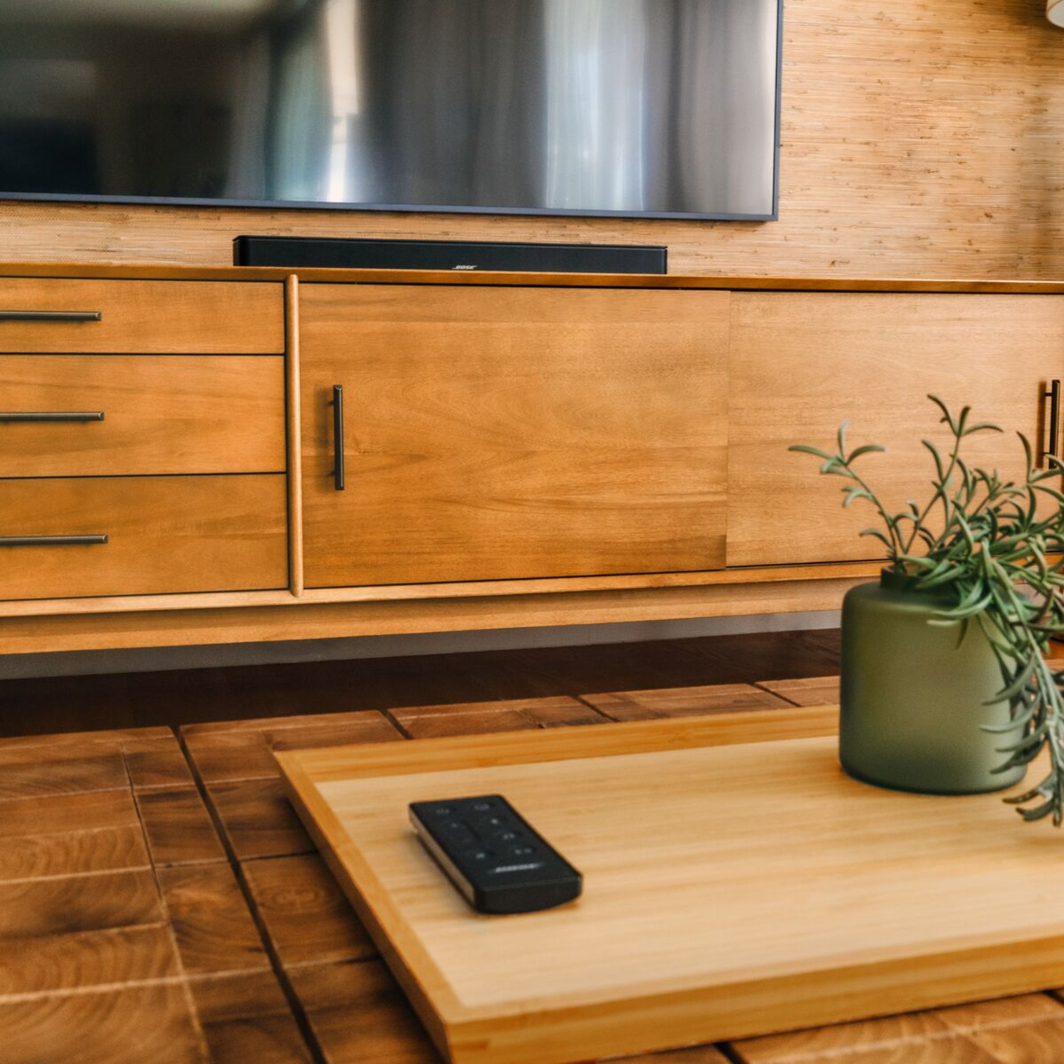 A wooden cabinet with a TV, a black soundbar, a tray with a remote, and a green vase with plants.