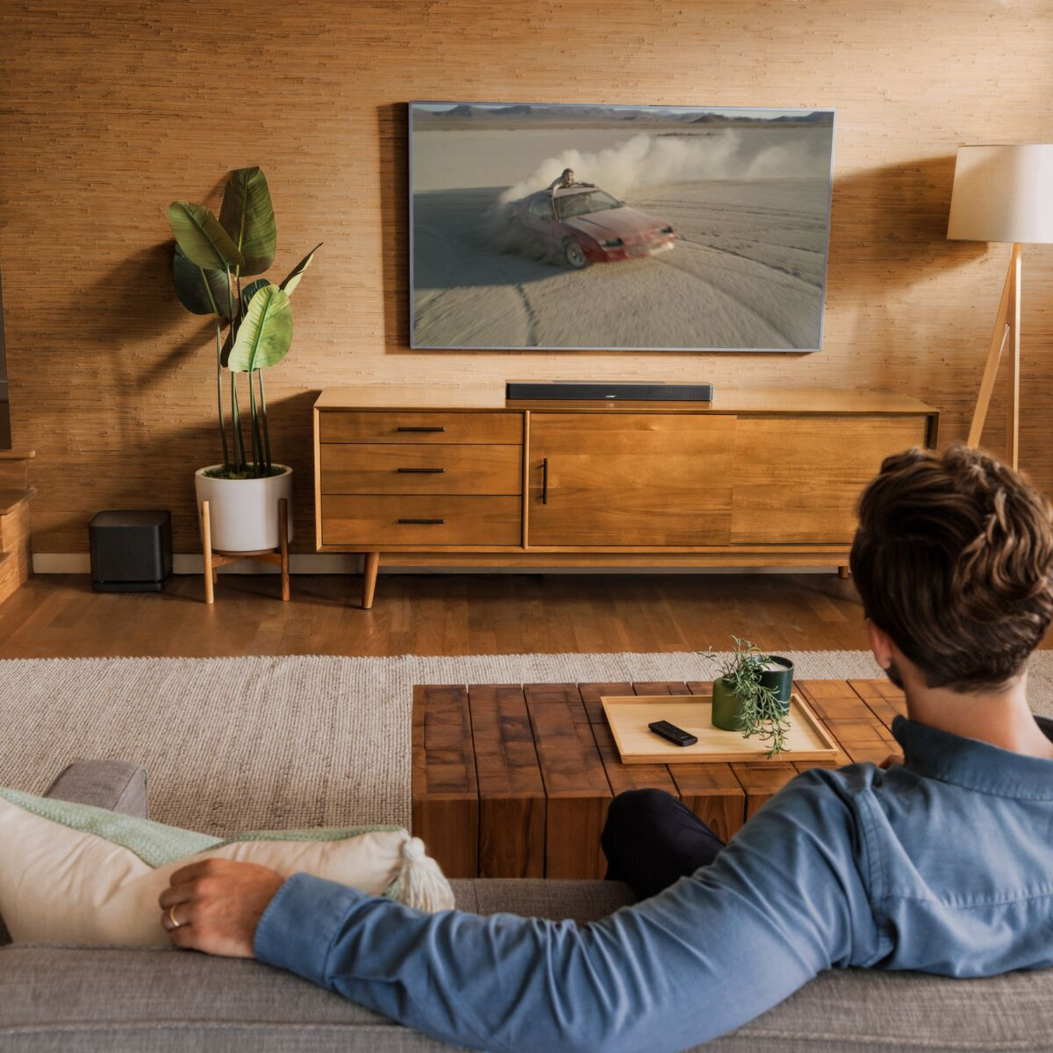 Living room scene. A person on a sofa watches TV above a wooden cabinet. Soundbar is visible.