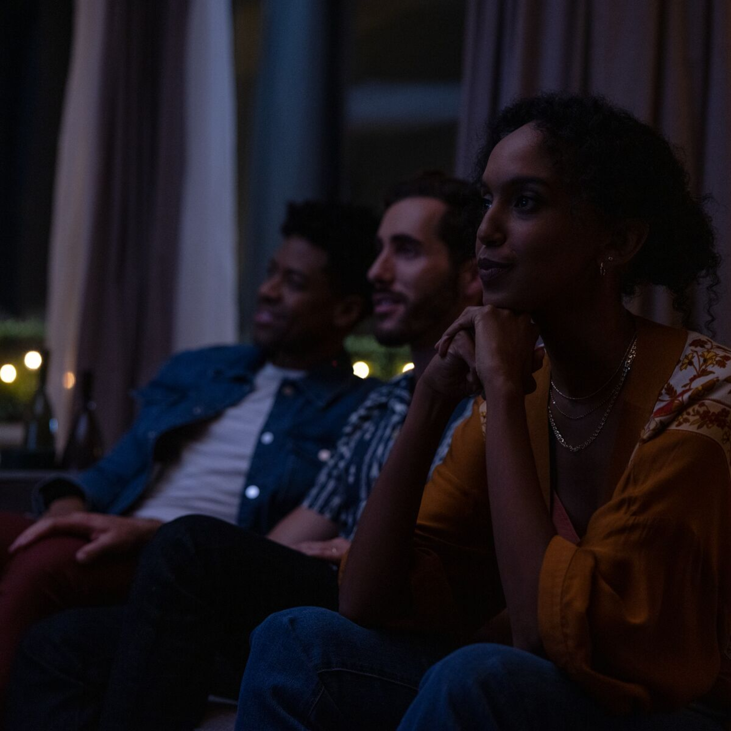 Three people watching something on a screen in a dark room, illuminated by a few lights in the background.