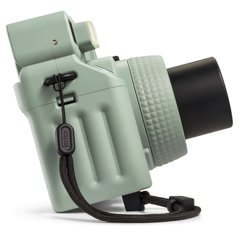 Zöld Instax kamera, ferde nézet. Fekete szíj és részletek. Fekete lencse. Fehér háttér.