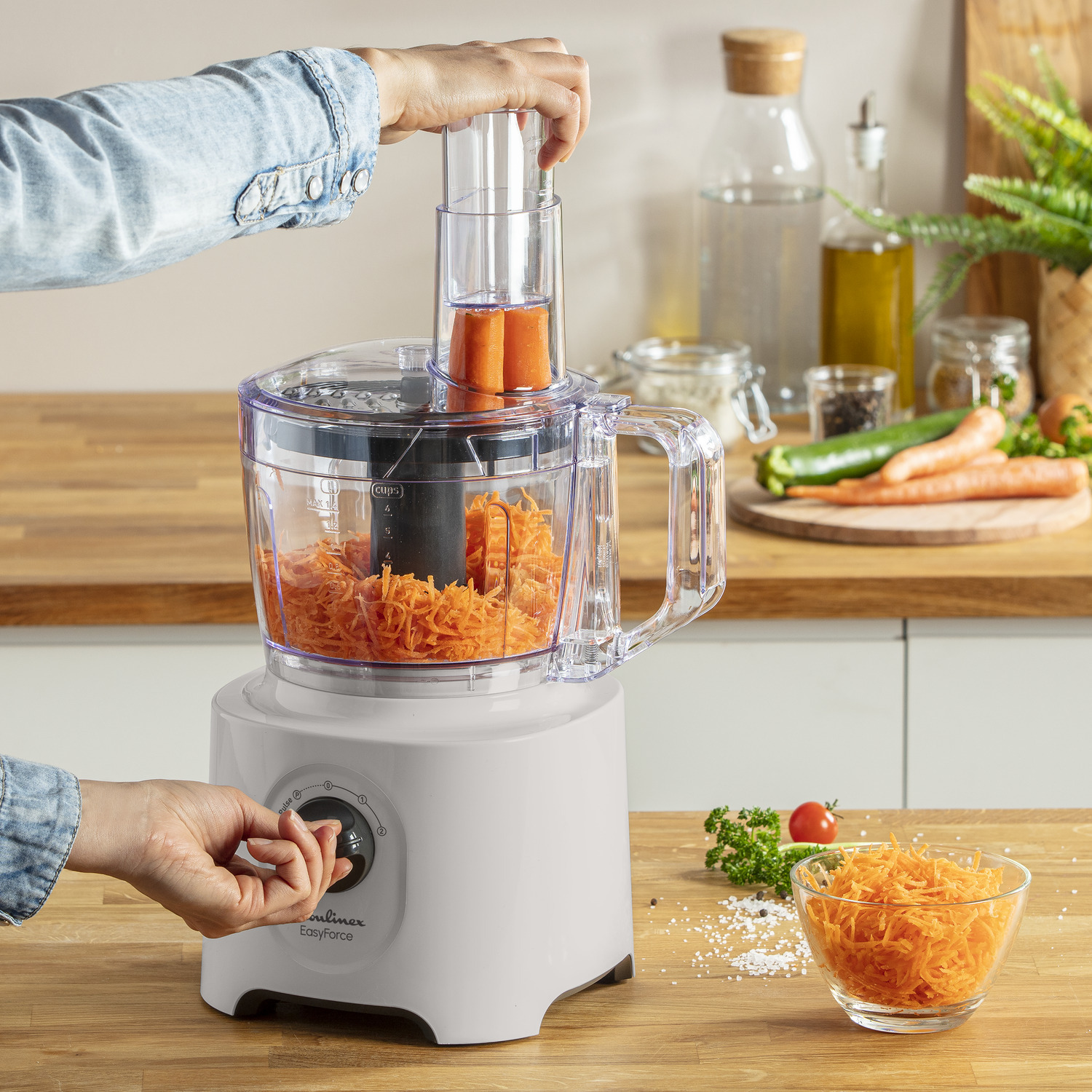 A food processor with a person adding carrots. Grated carrots in the bowl.