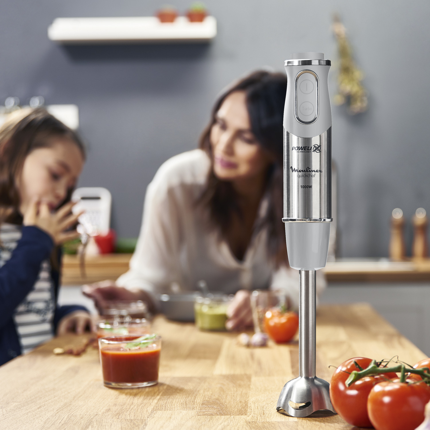 A woman and child near a table with a hand blender mixing tomato sauce, and ingredients.