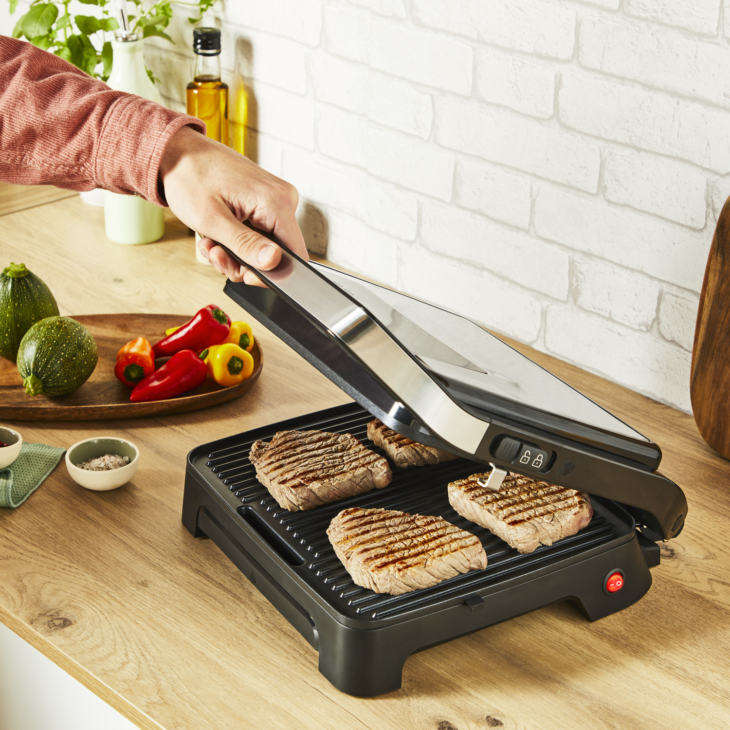 Person grilling steaks on an indoor grill with vegetables nearby.