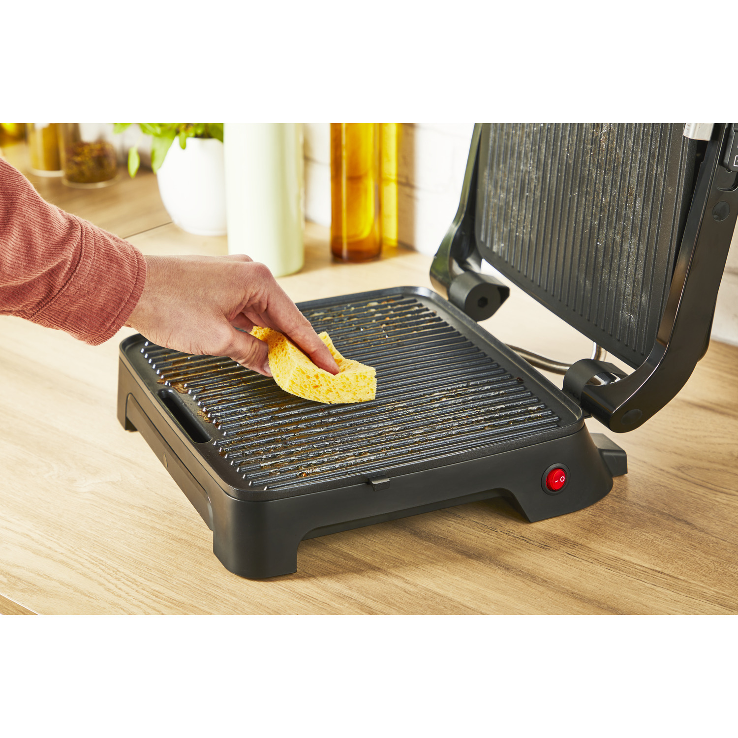 A hand cleaning a dirty black grill with a yellow sponge, on a wooden surface.