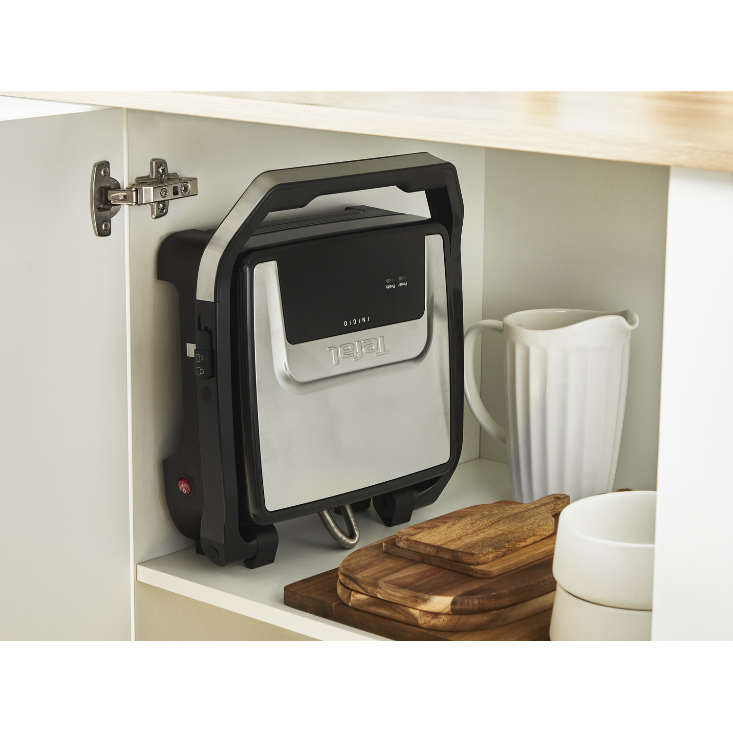 A black and silver Tefal Inicio grill stored in a white cabinet with other kitchen items.