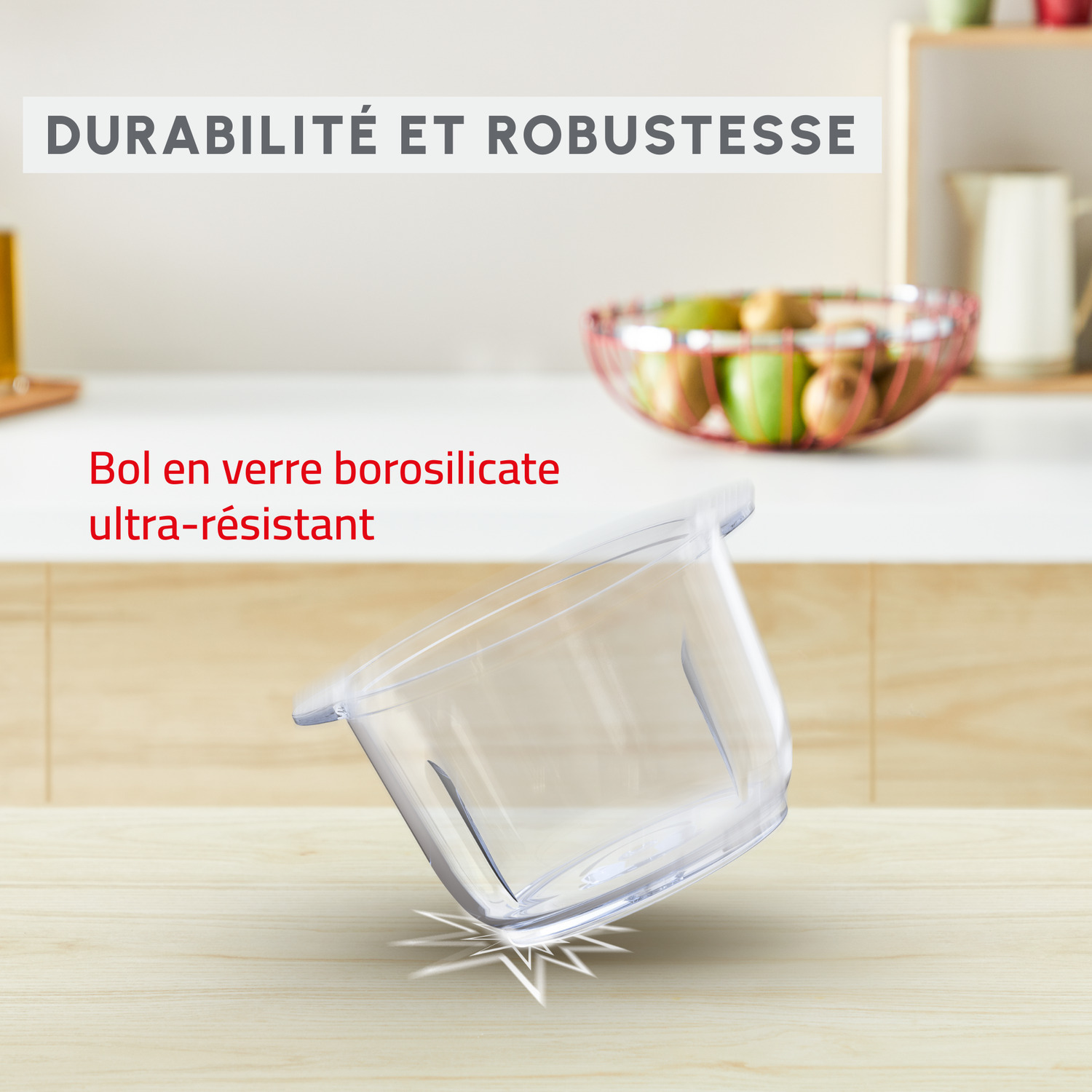 Bol en verre transparent incliné sur une table en bois, avec un comptoir blanc et un panier de fruits en arrière-plan.