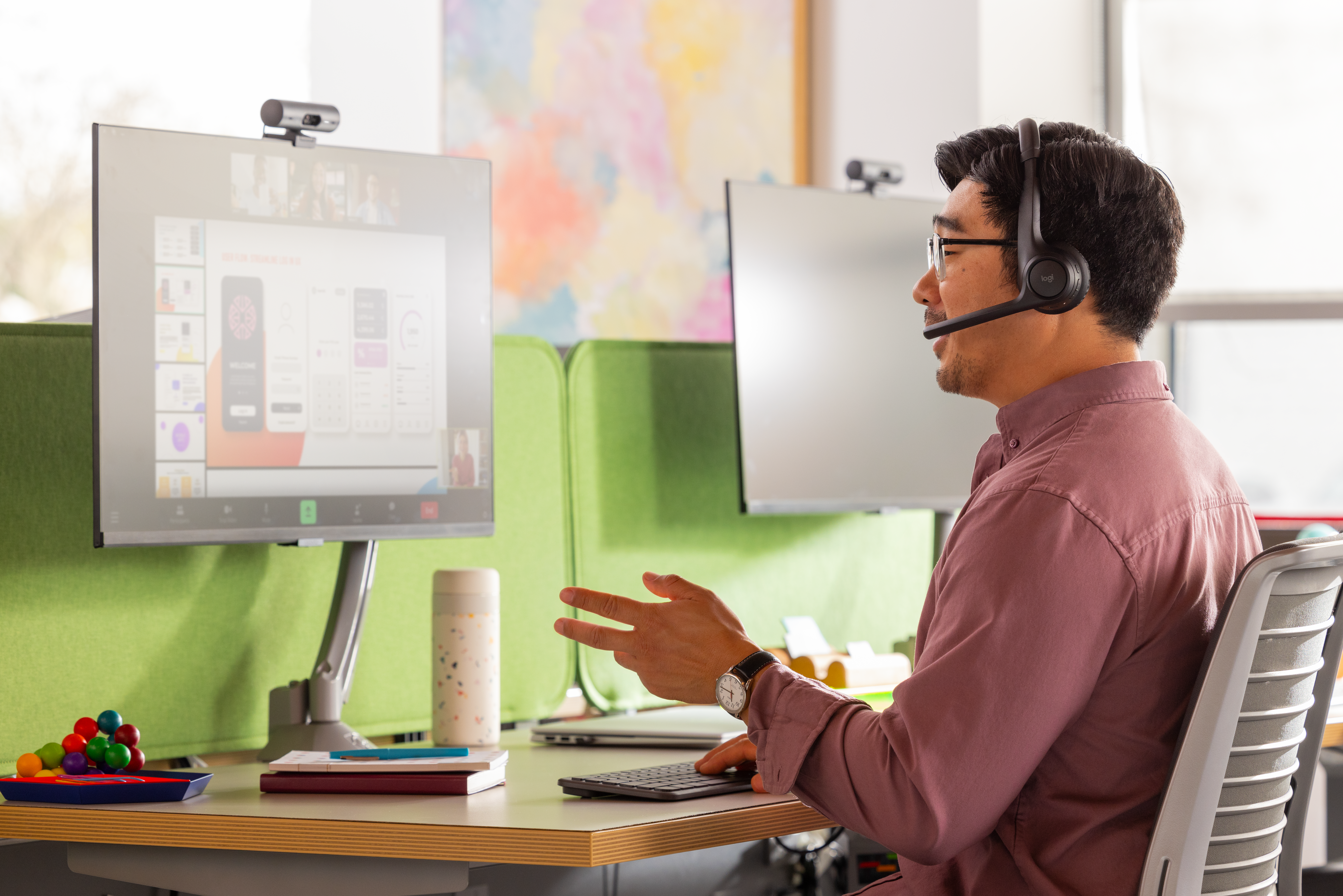 Mann im Meeting mit Headset. Zwei Bildschirme zeigen Videoanruf, Gegenstände auf dem Tisch.