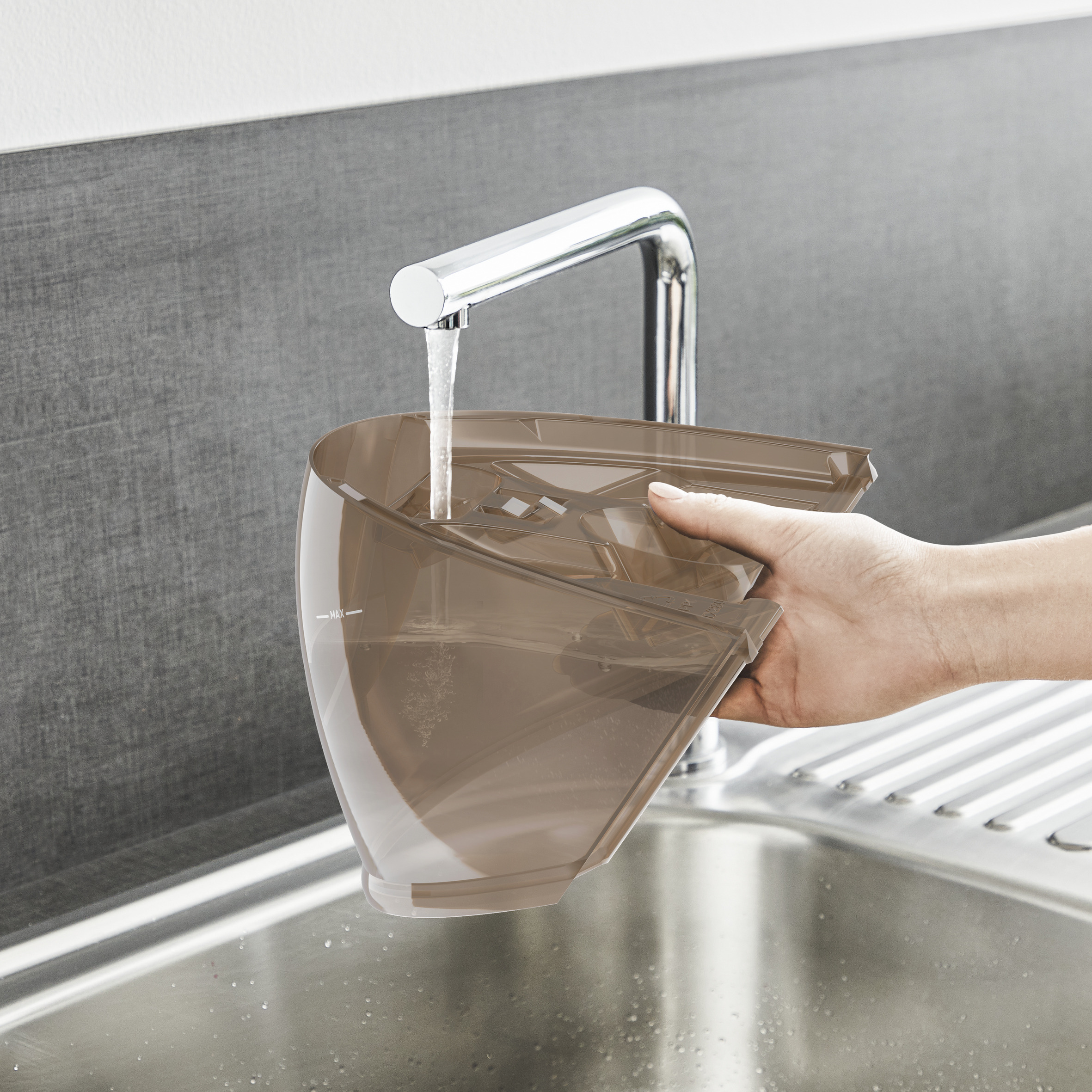Ręka napełniająca brązowy pojemnik wodą w zlewie. Chromowana bateria, szary zlew, białe tło.