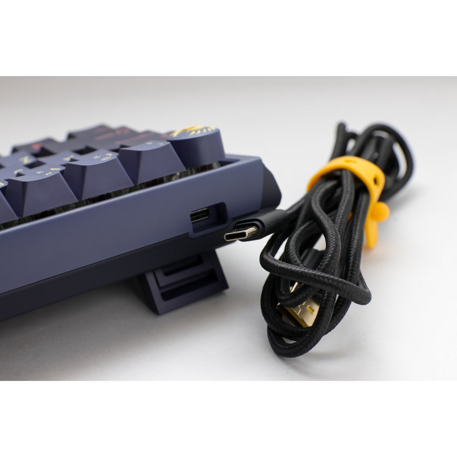 Un teclado mecánico azul con cable USB-C negro y brida amarilla. Fondo blanco.