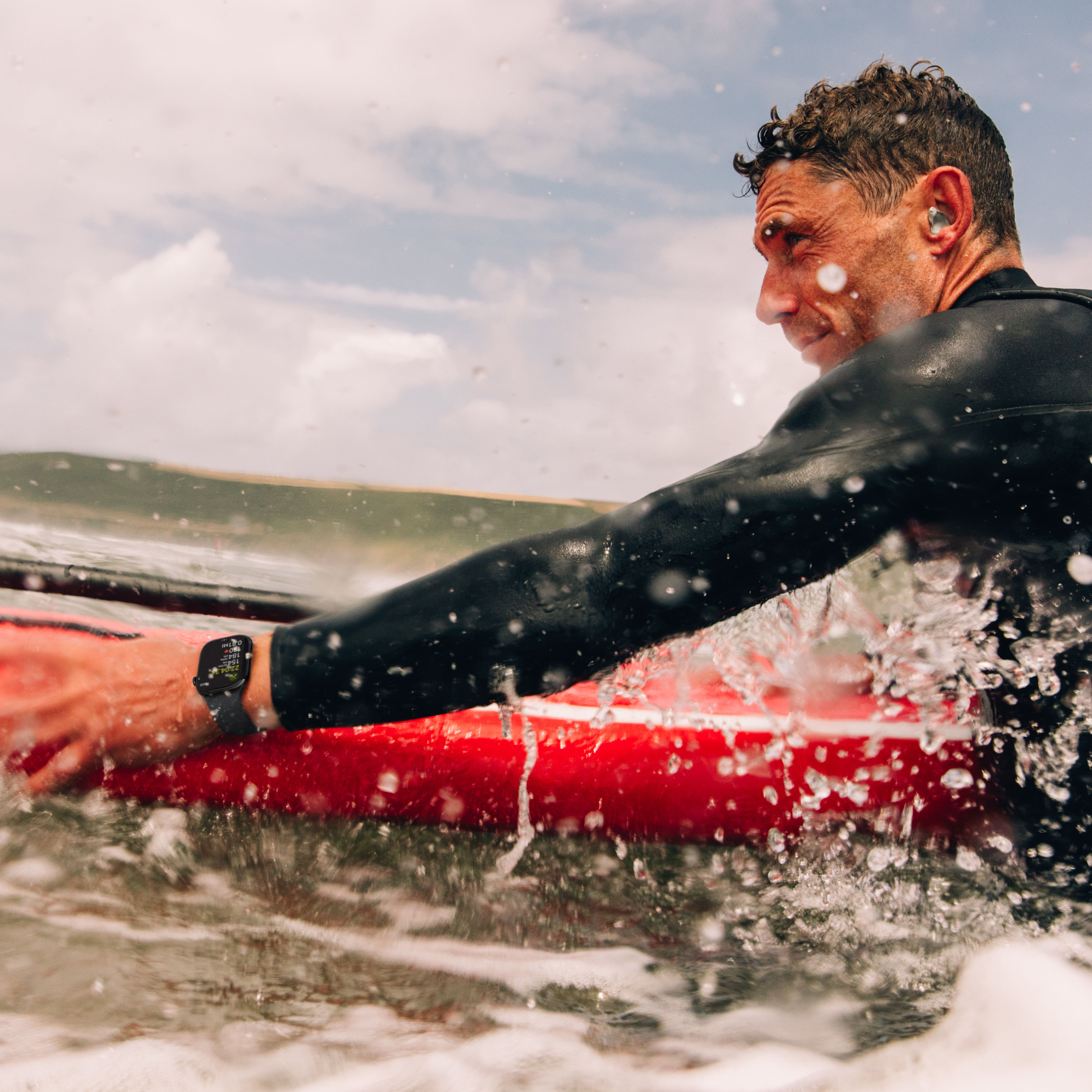 Un homme surfant avec une Apple Watch au poignet, de l'eau éclaboussant autour. Ciel nuageux.
