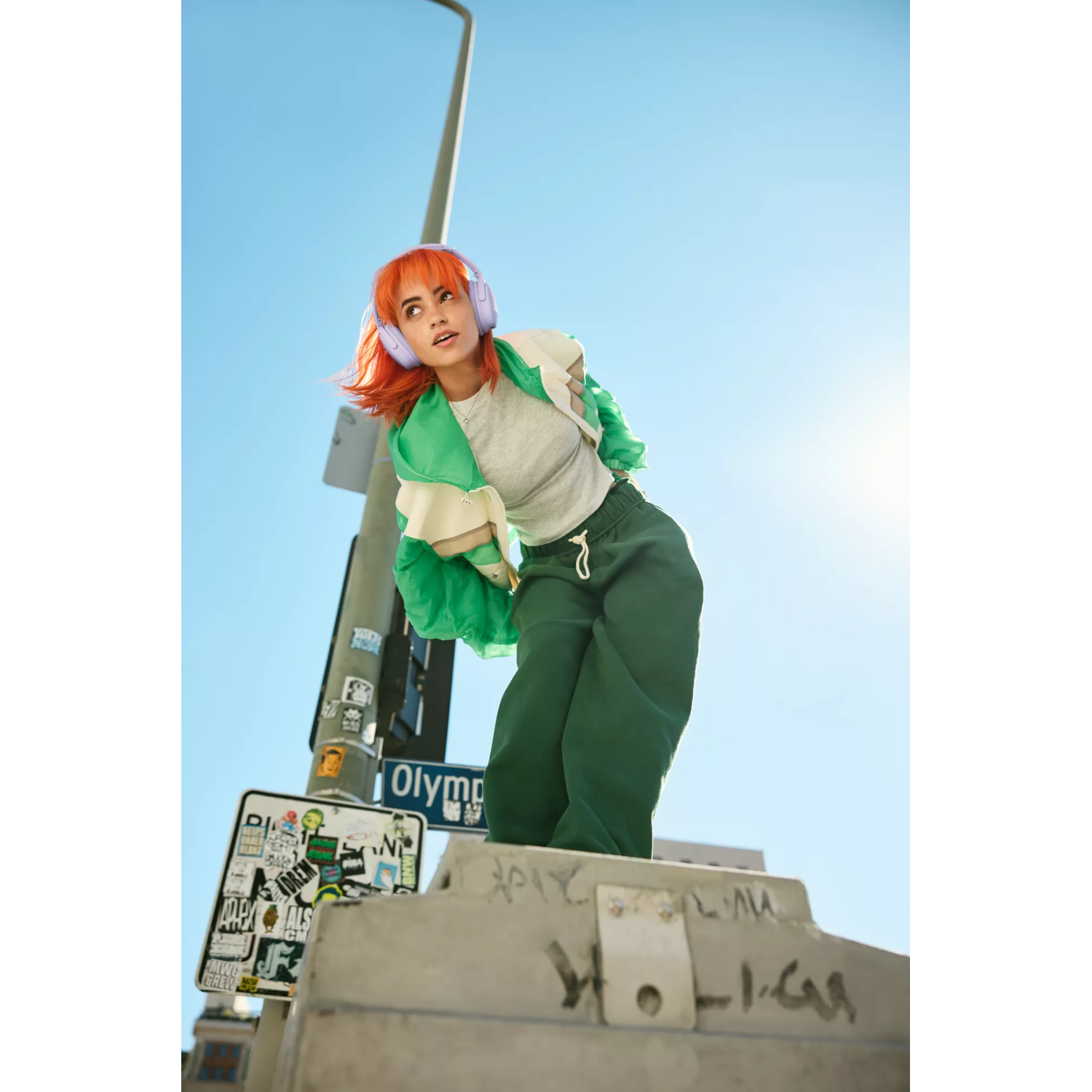 A woman with orange hair wears headphones, standing on a street sign, blue sky background.