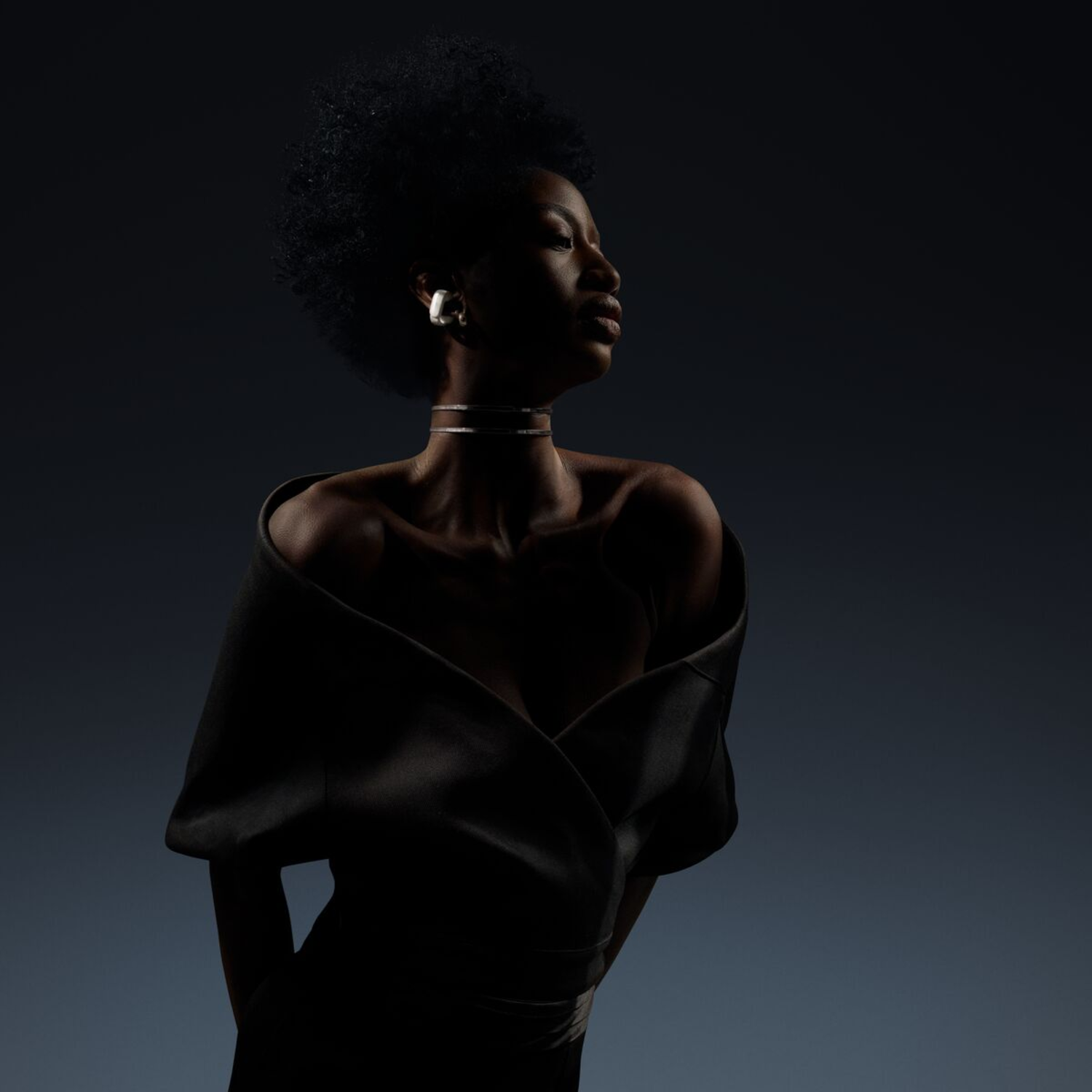 A woman in a black dress, with silver earrings, looking away, in a dark studio setting.