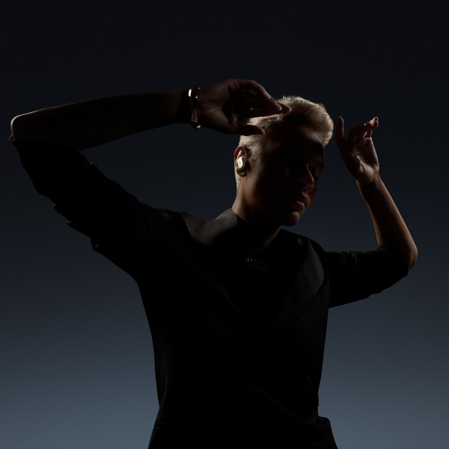 Silhouette of a person with blonde hair, wearing earbuds, hands up, dark background.