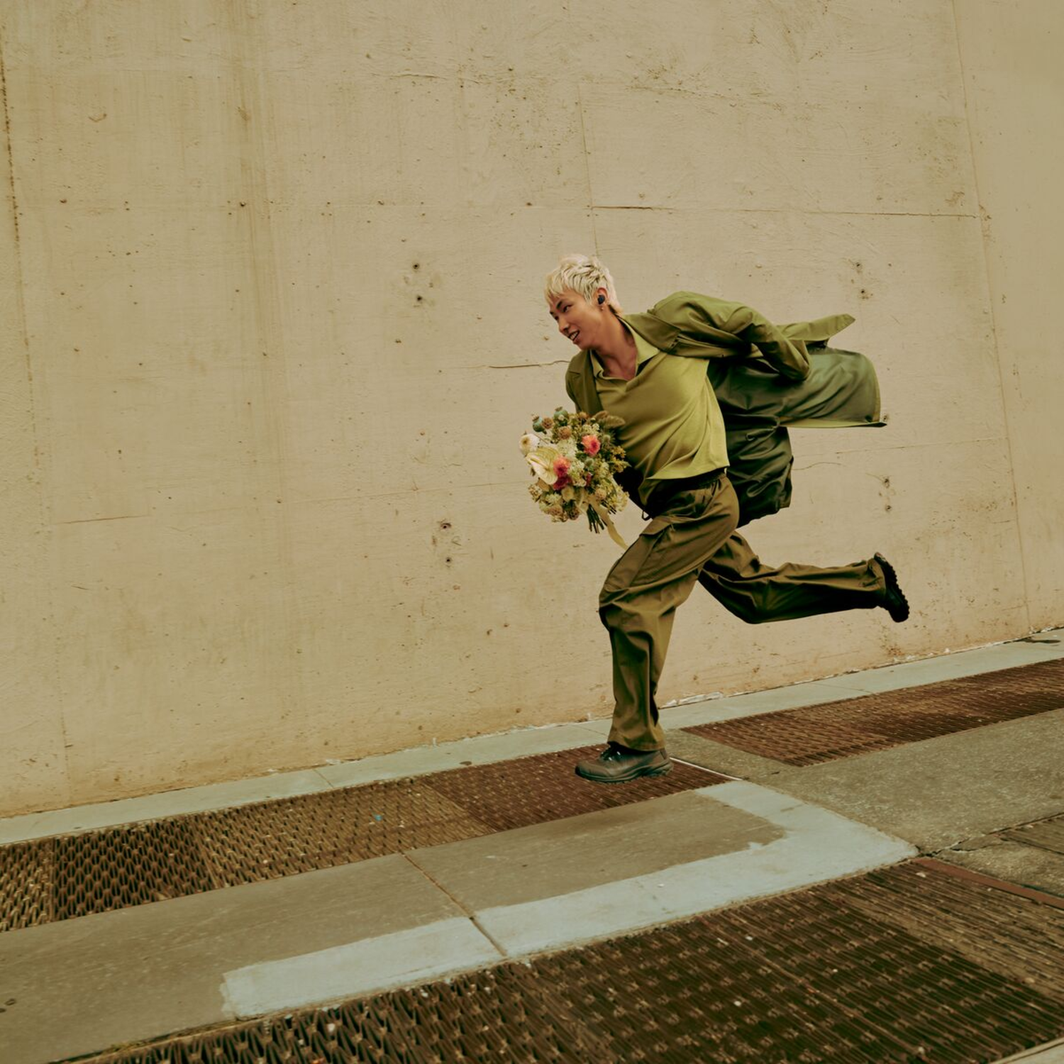 A man in green clothes running with flowers, next to a concrete wall.
