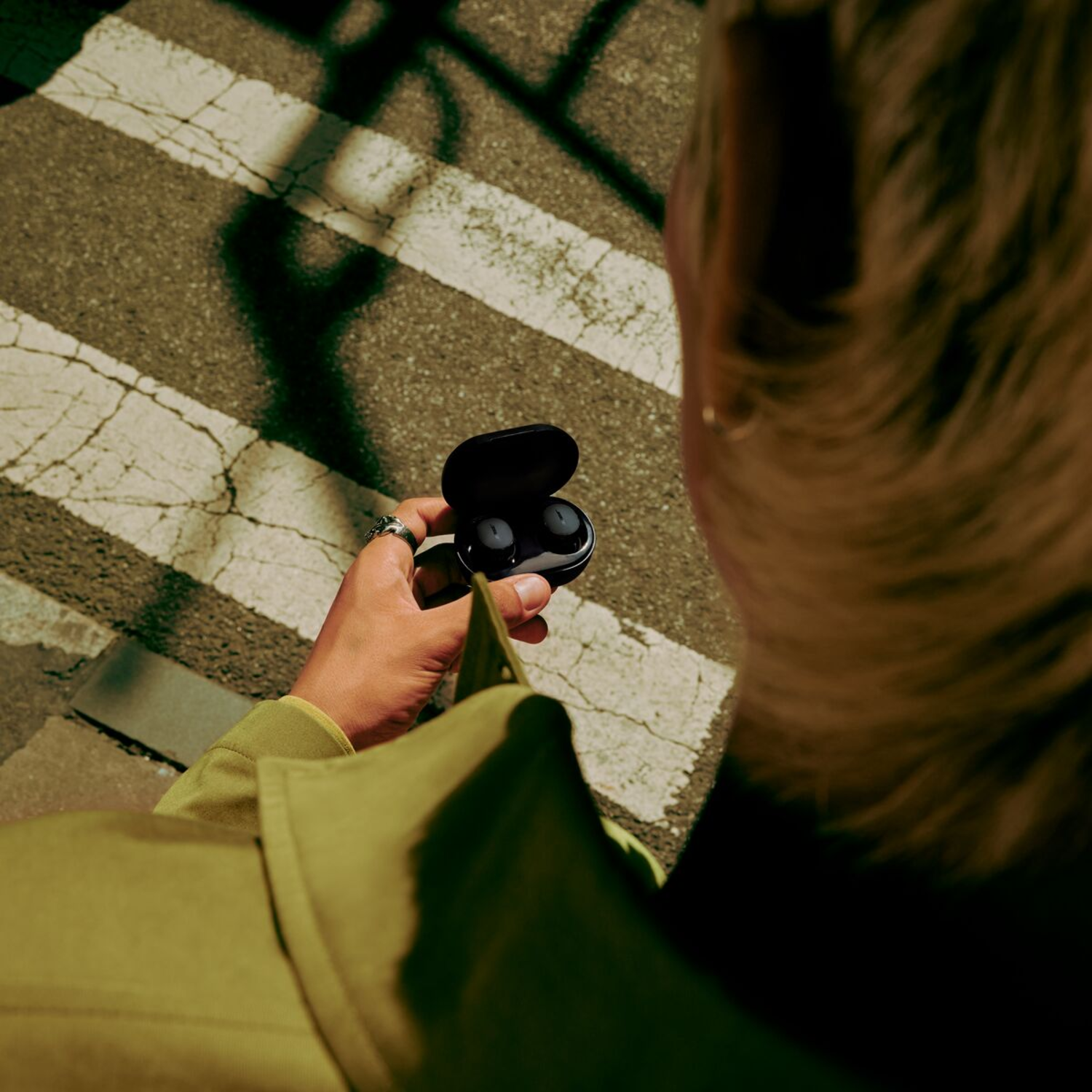 Person holding a black earbuds case, with earbuds visible. They are in a public setting.