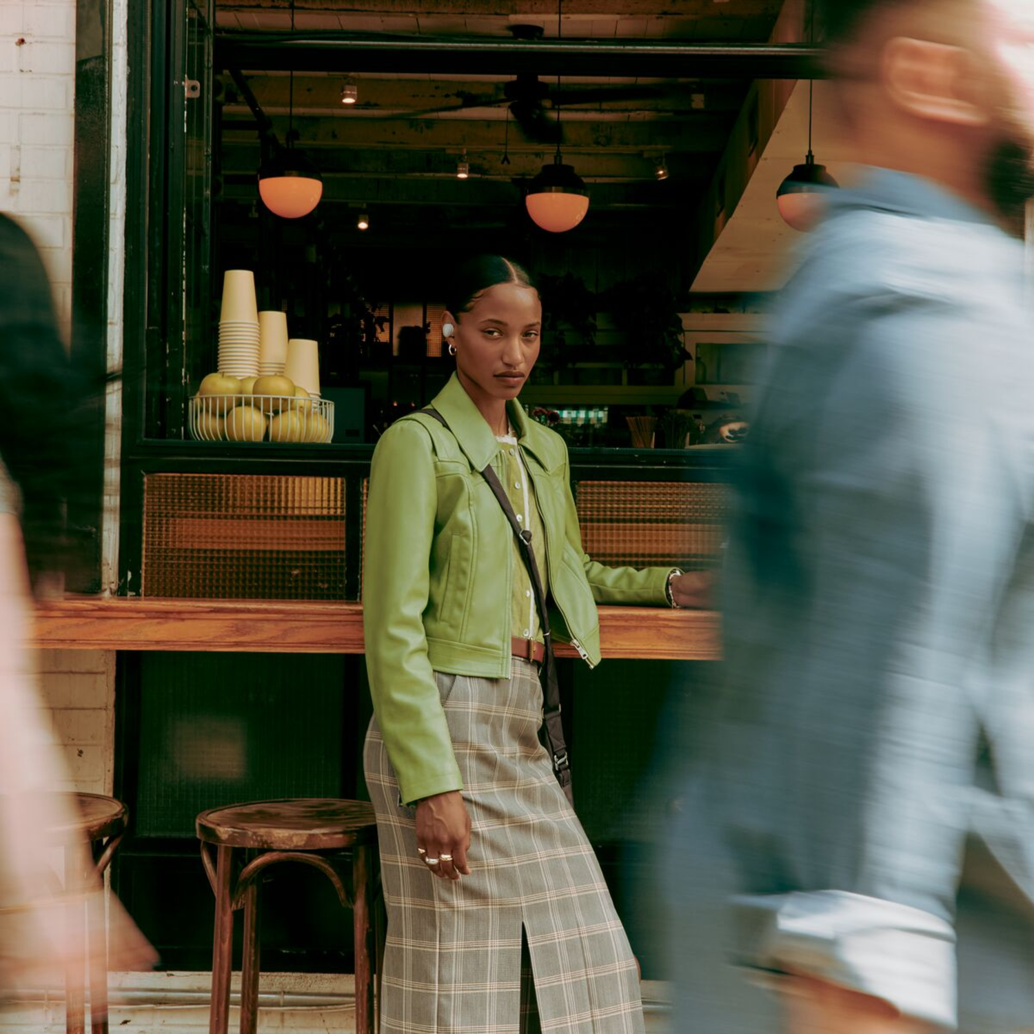 Woman in green jacket and skirt standing outside a cafe with earbuds in her ears.
