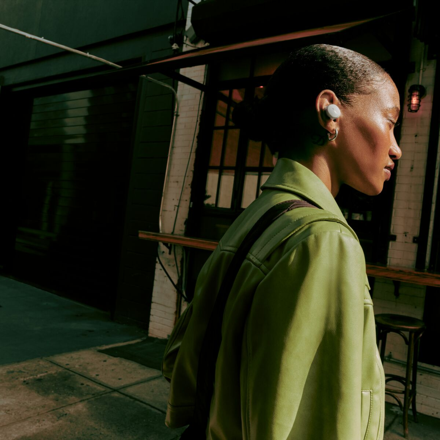 Woman with earbuds, green jacket, walking on a street, near a brick building with a sign.