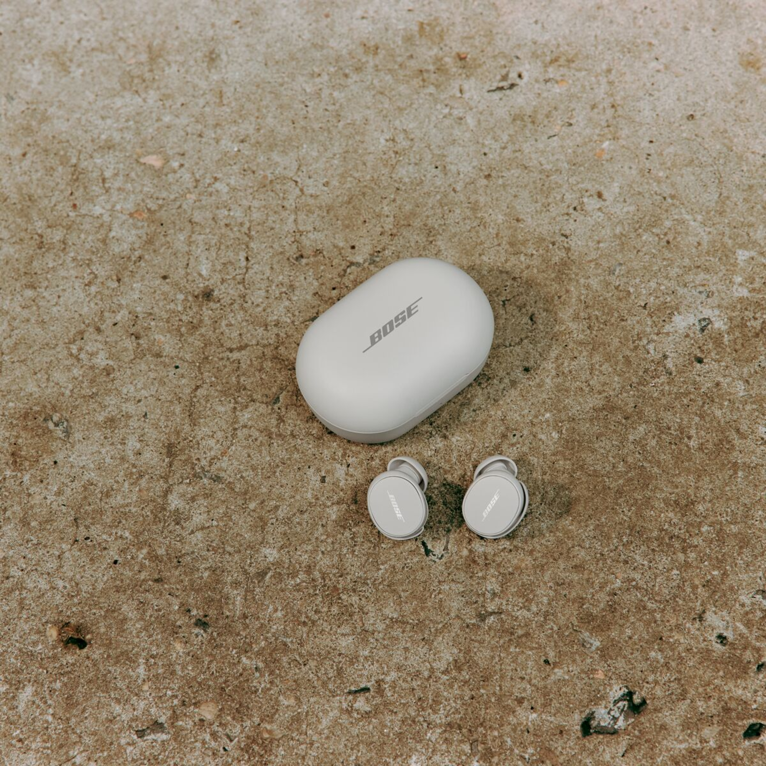 White Bose earbuds and charging case on a textured concrete surface. Top-down view.
