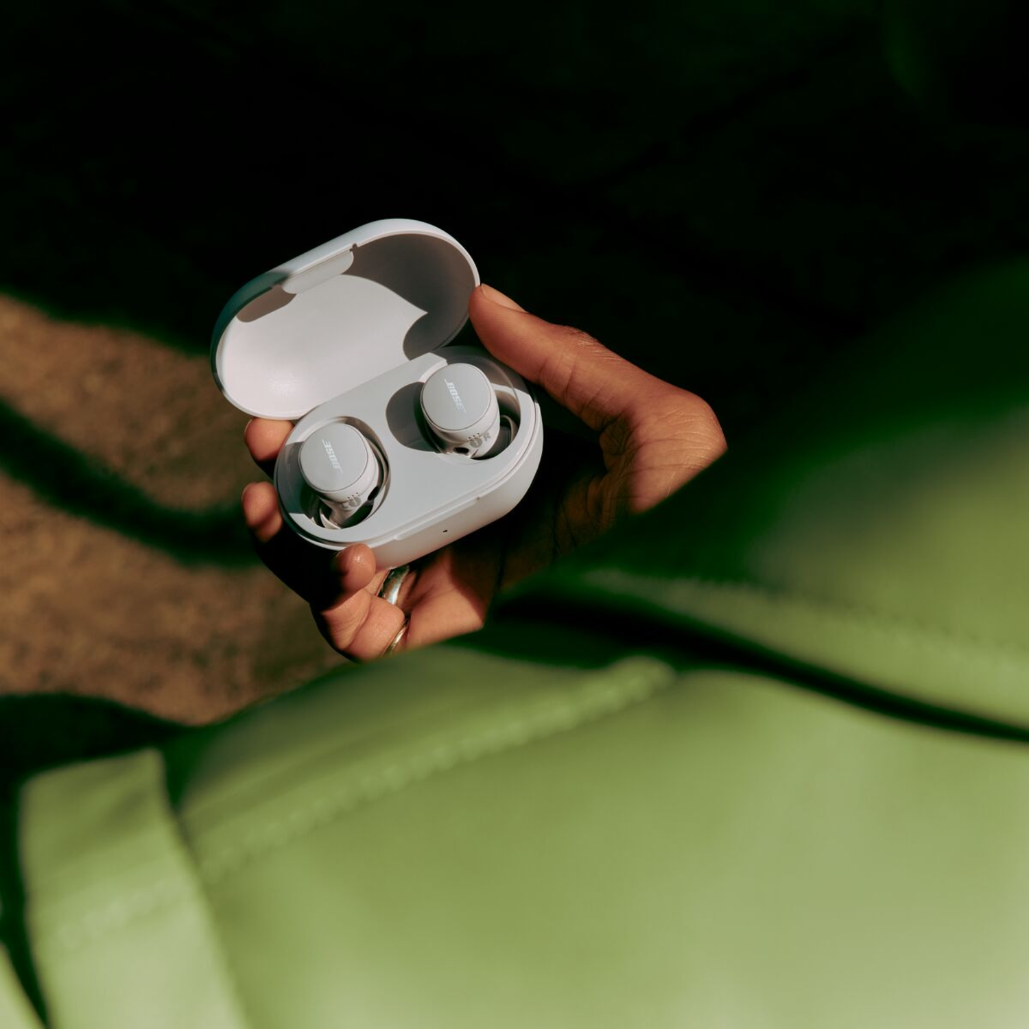 A hand holding a white open case with two gray earbuds. The background is blurred green.