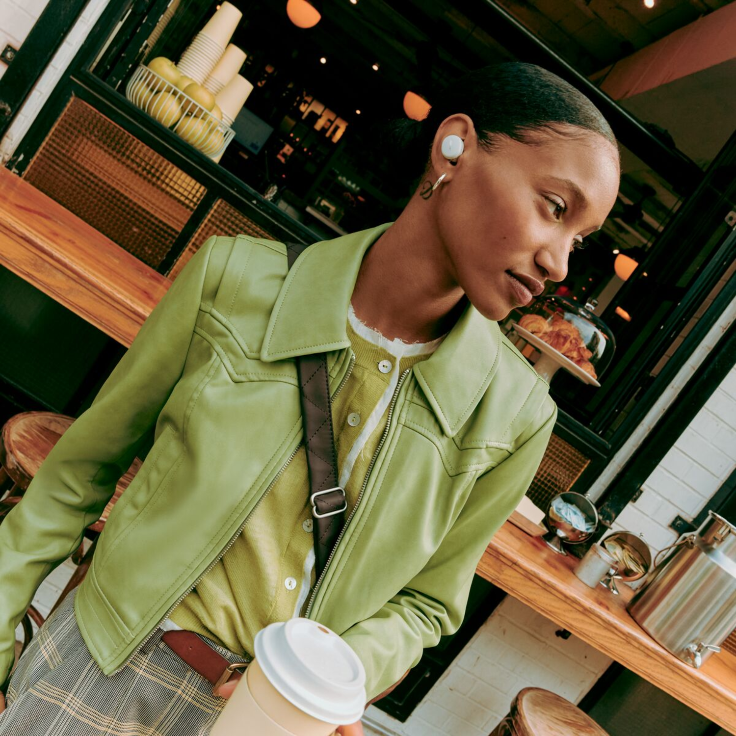 A woman in a green jacket and earrings holds a coffee cup near a cafe with a wooden counter.