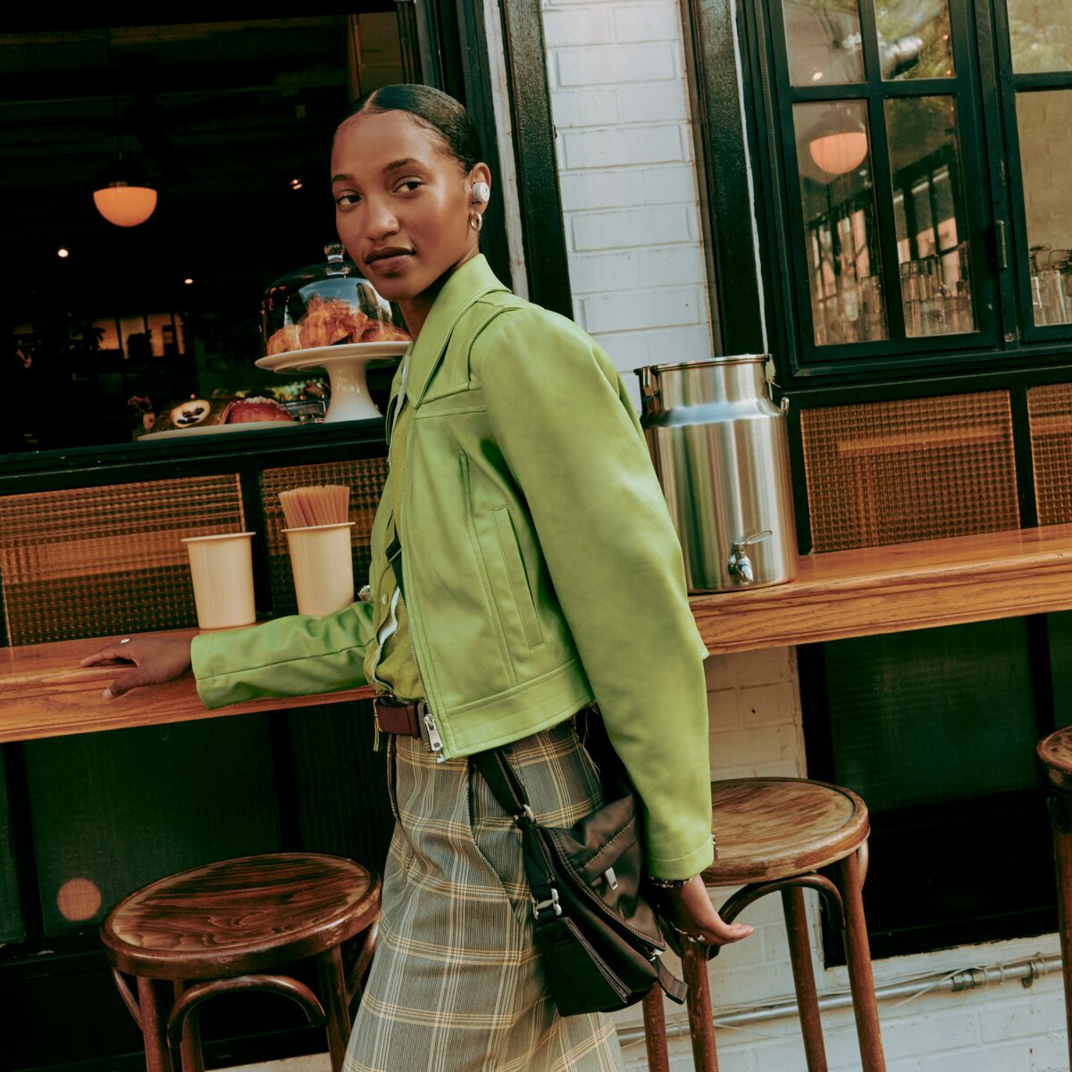 Woman in green jacket walking, wearing earbuds, in front of a shop with a wooden counter.