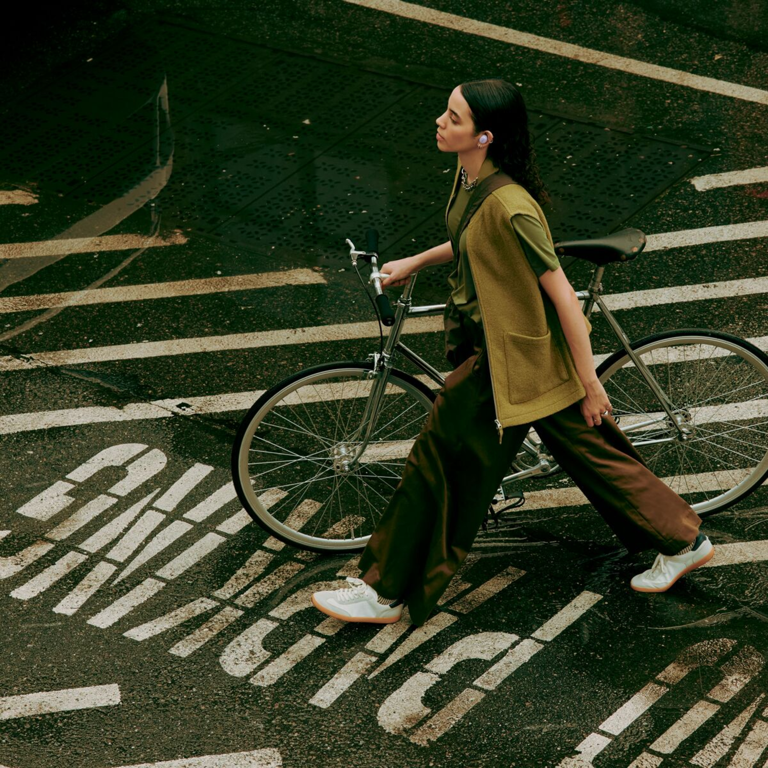Vrouw met koptelefoon die op de fiets loopt. De scène is een straat met nat wegdek.