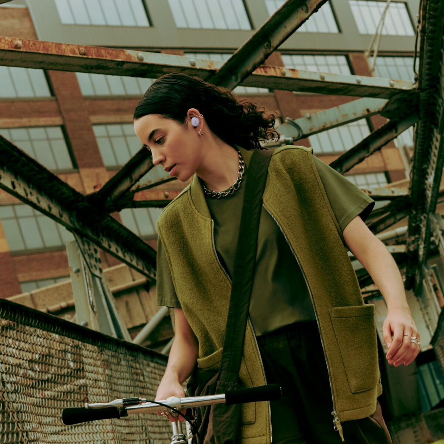 Vrouw met oordopjes fietst op een brug, met een groen vest en jas.