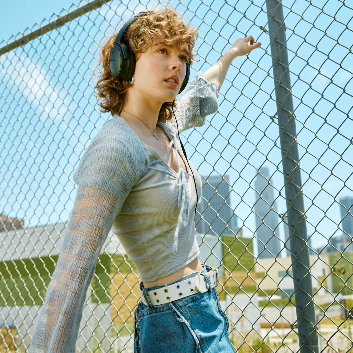 Woman with headphones by a fence, looking up at a city skyline.