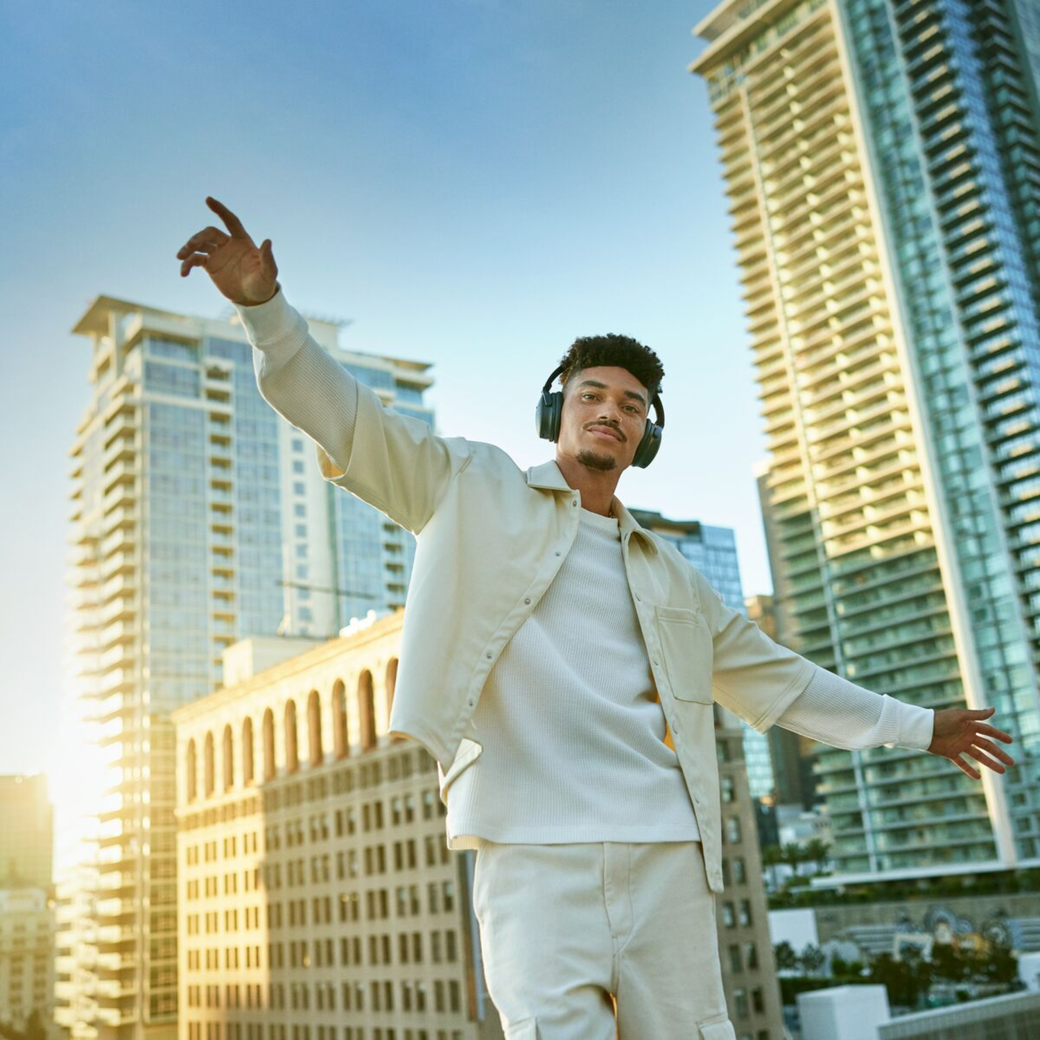 A man wearing headphones on a rooftop in a city with tall buildings, arms outstretched.