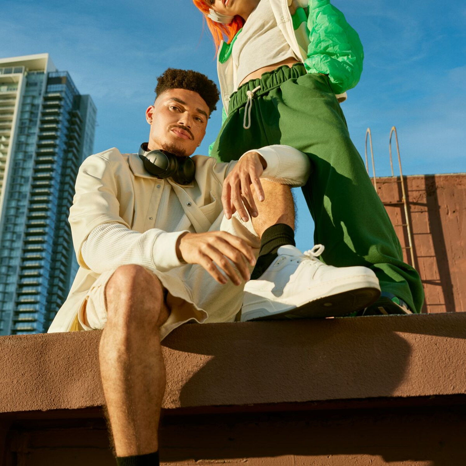 Two people sitting on a rooftop, with a cityscape and blue sky background.