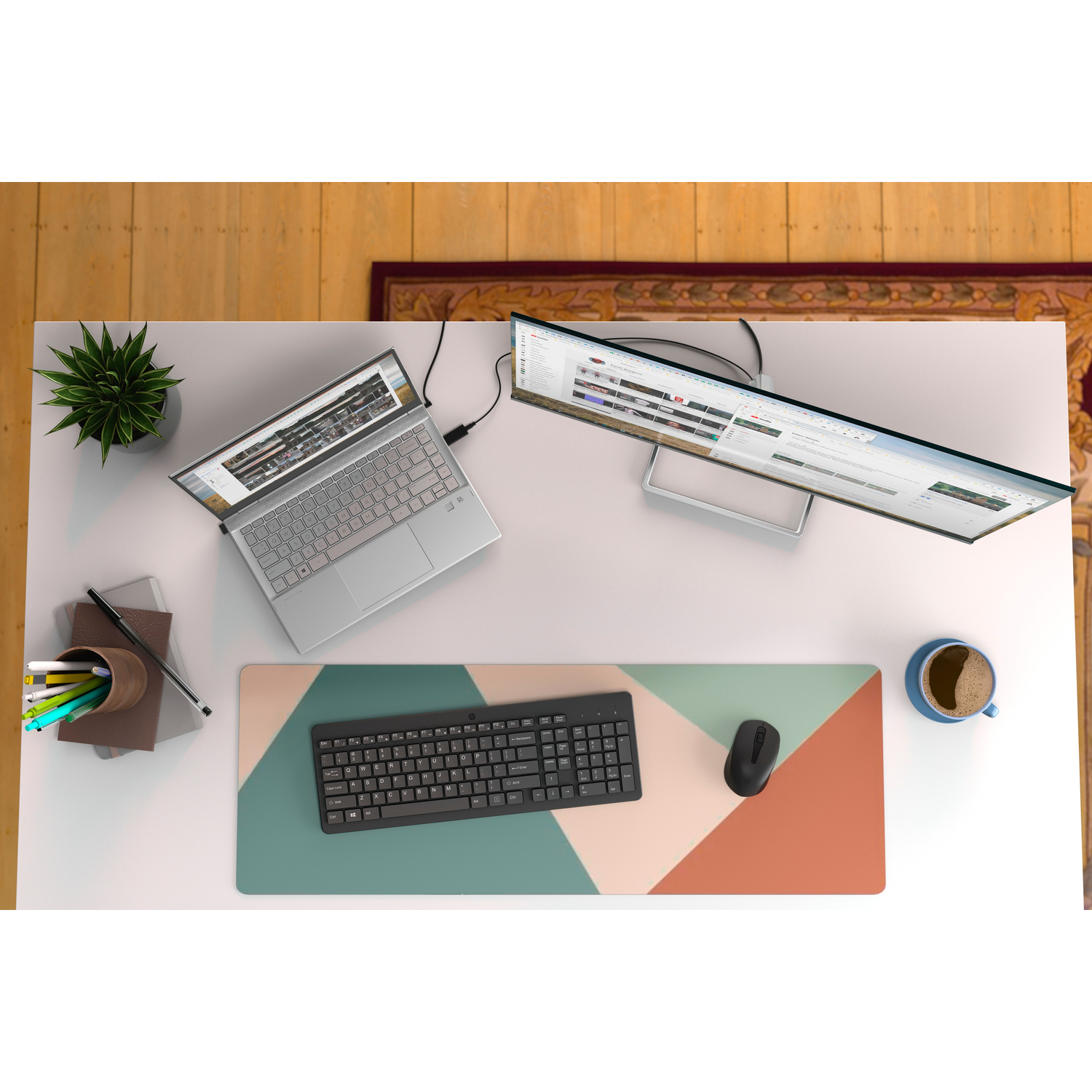 Bovenaanzicht van een bureau met een laptop, monitor, toetsenbord, muis, koffiekopje en plant.