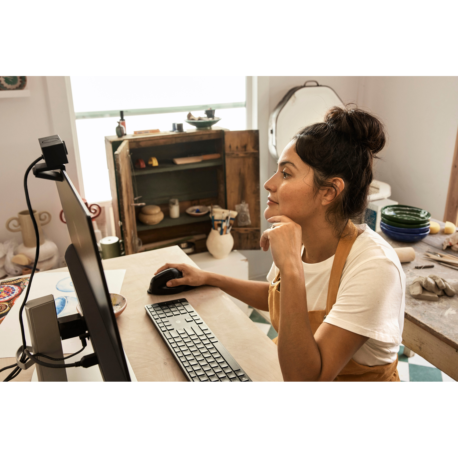 Woman using a computer, mouse in hand. Background: pottery, window, and wooden cabinet.