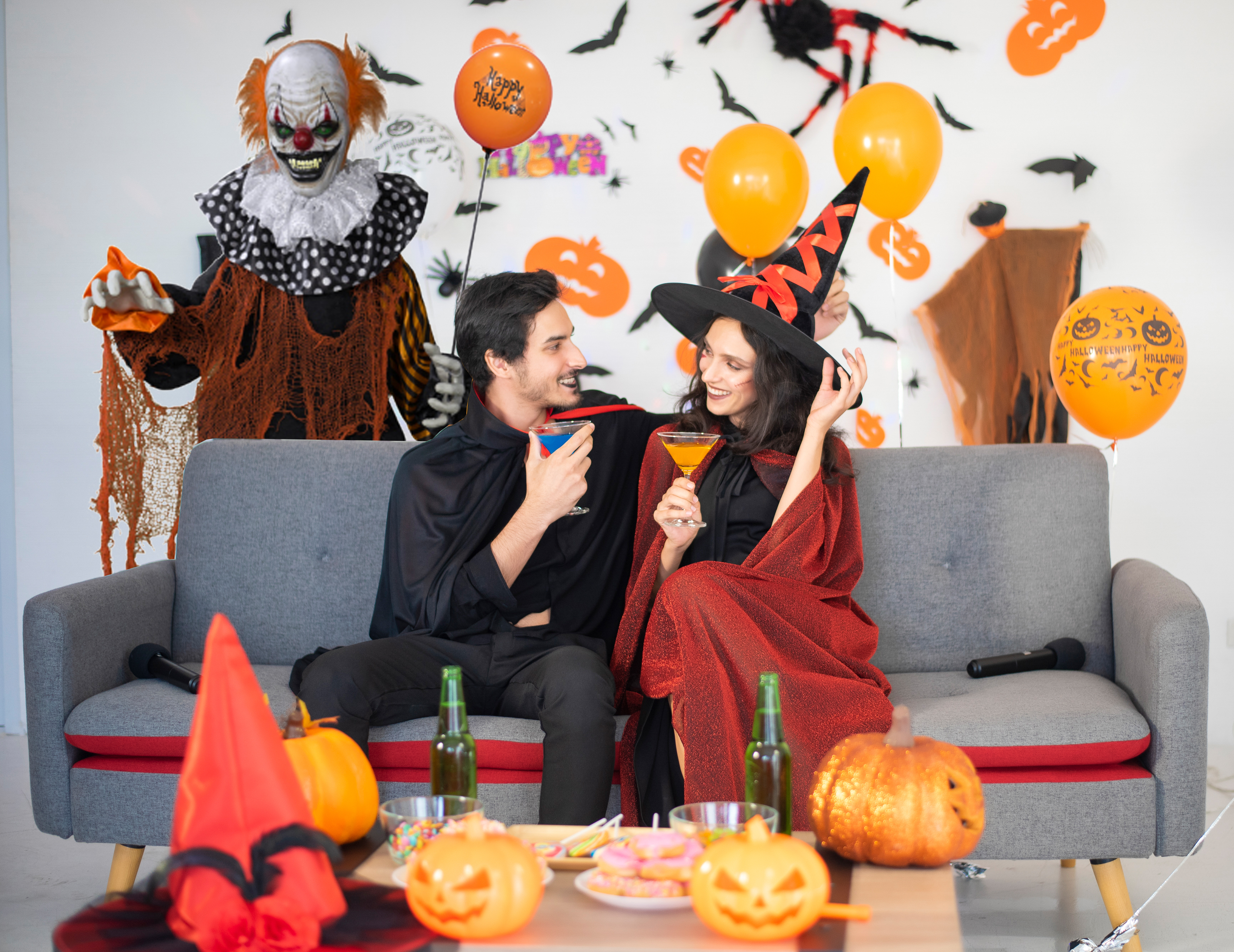 Ein Paar in Halloween-Kostümen sitzt mit Getränken auf einem Sofa, dahinter eine Clown-Requisite und Kürbisse.