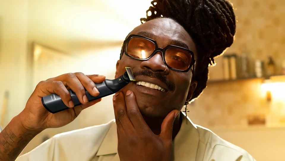 Mężczyzna goli twarz trymerem, nosząc okulary i uśmiechając się. Łazienka jest w tle.