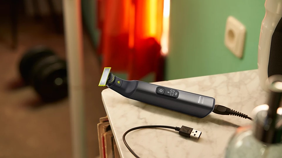 Trymer elektryczny ładujący się na marmurowej powierzchni. Kabel USB podłączony. Tło z zieloną ścianą i pomarańczowym akcentem.