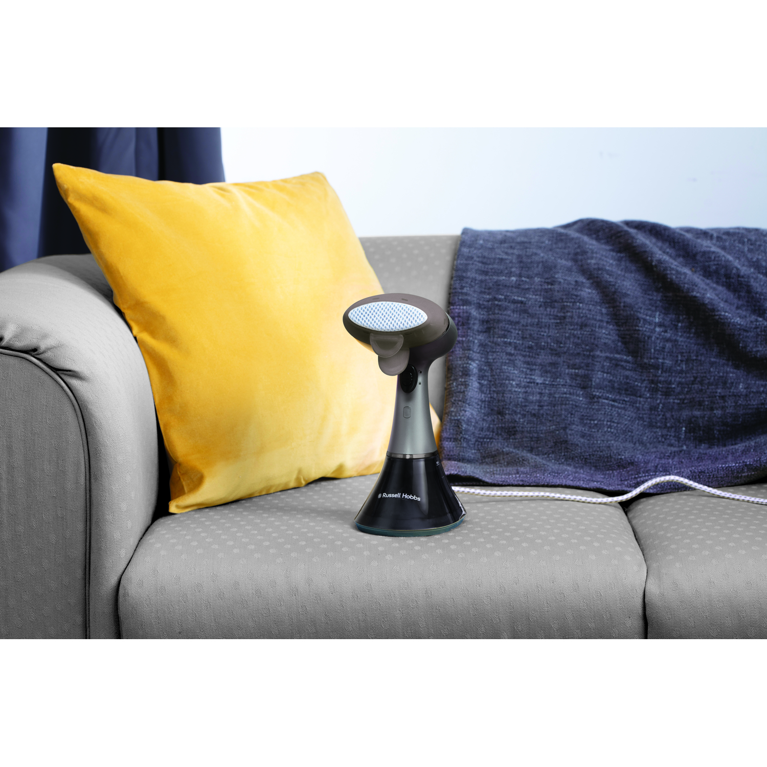 A handheld clothes steamer on a grey sofa with yellow pillow and grey blanket.