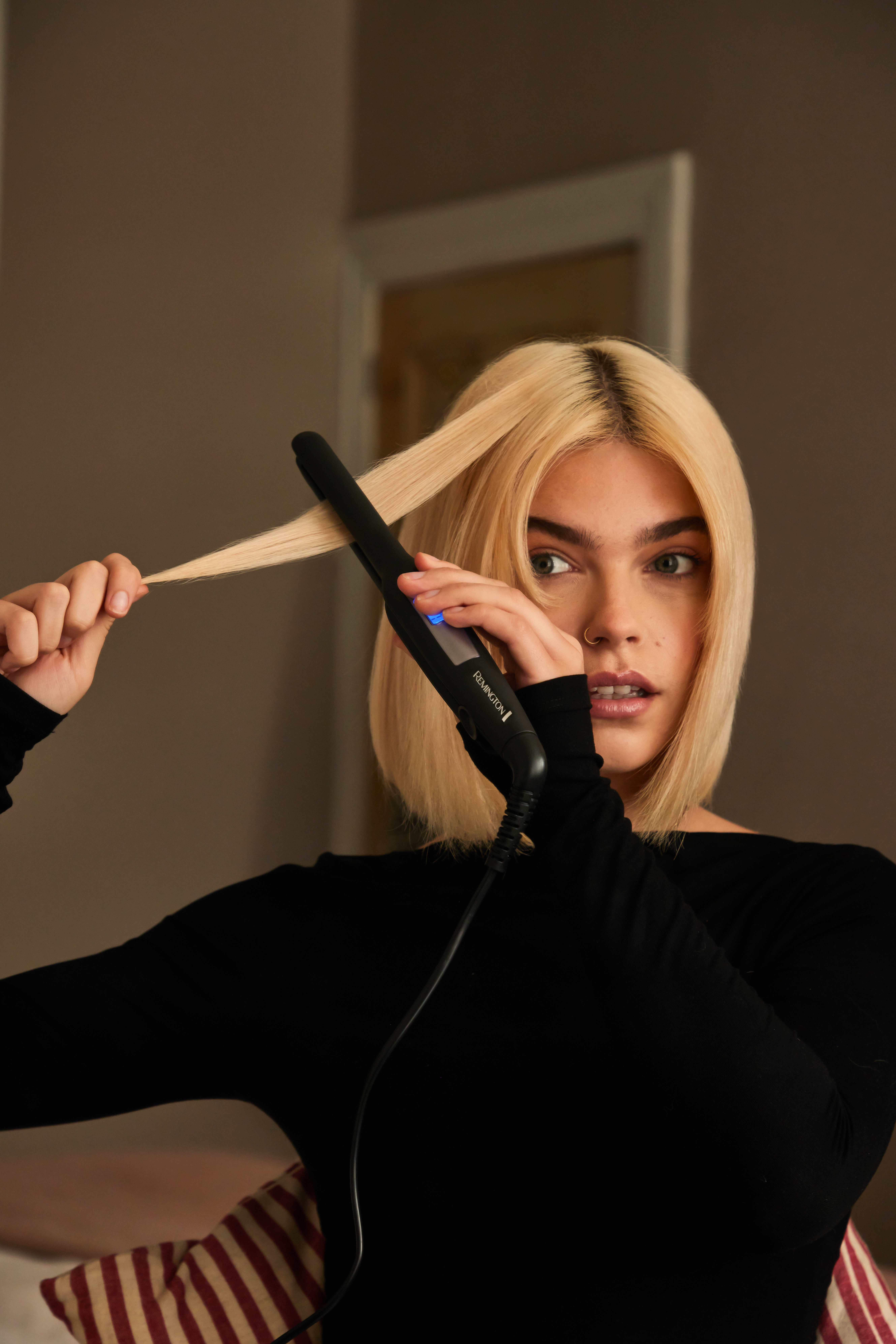 Eine Frau mit blonden Haaren benutzt ein schwarzes Glätteisen. Sie trägt ein schwarzes Oberteil.