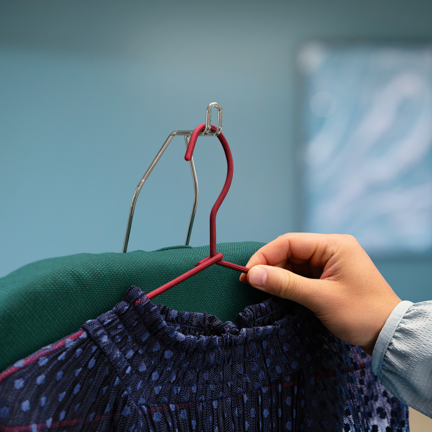 Hand die een donkerrode kleerhanger vasthoudt, met een groen overhemd en een blauw overhemd. Blauwe muur achtergrond.
