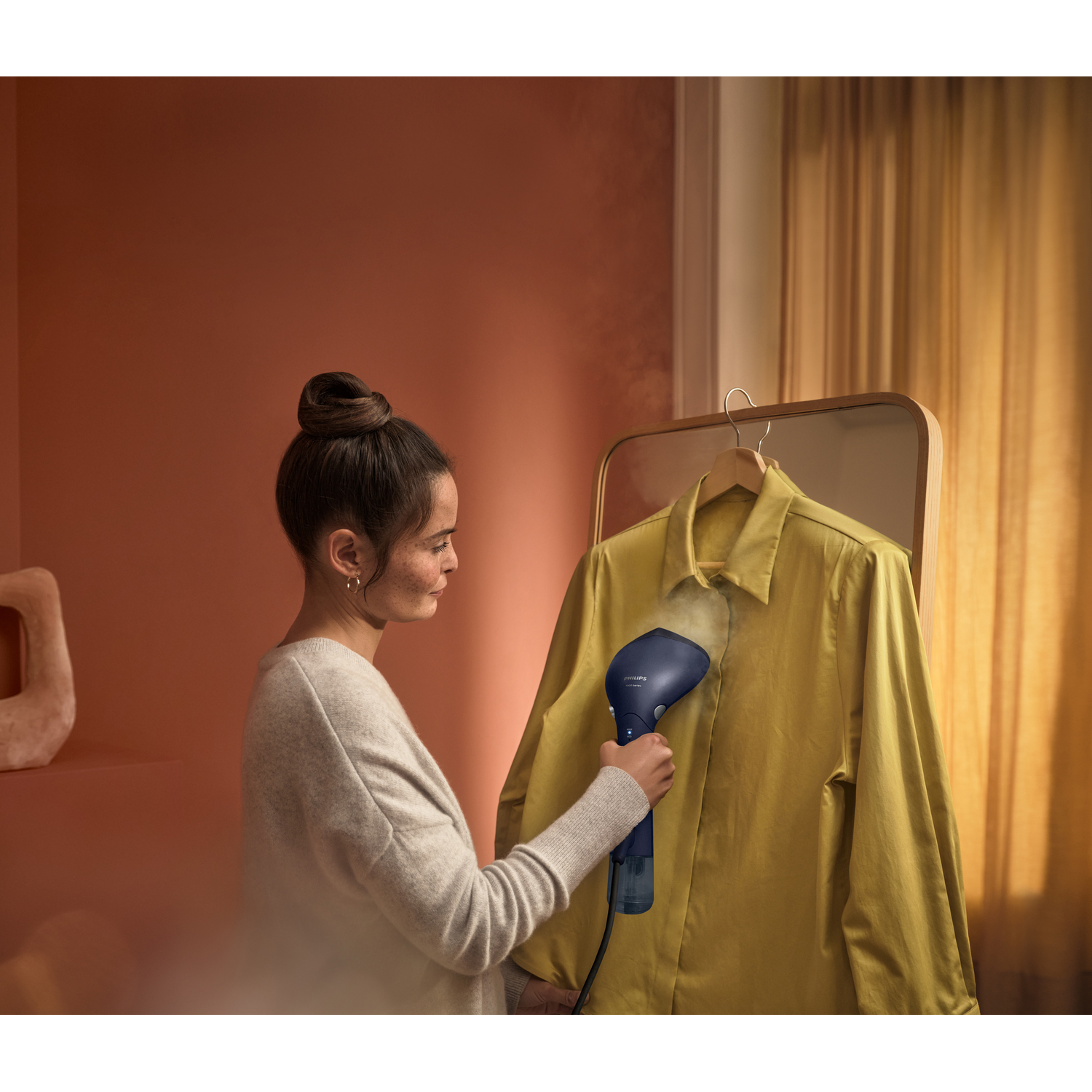 Vrouw die een geel overhemd stoomt met een blauwe Philips stomer. Een spiegel en gordijn op de achtergrond.