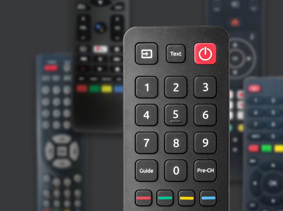 TV-Fernbedienung. Schwarz mit Zahlen, Text und Ein-/Aus-Taste. Andere Fernbedienungen im Hintergrund.