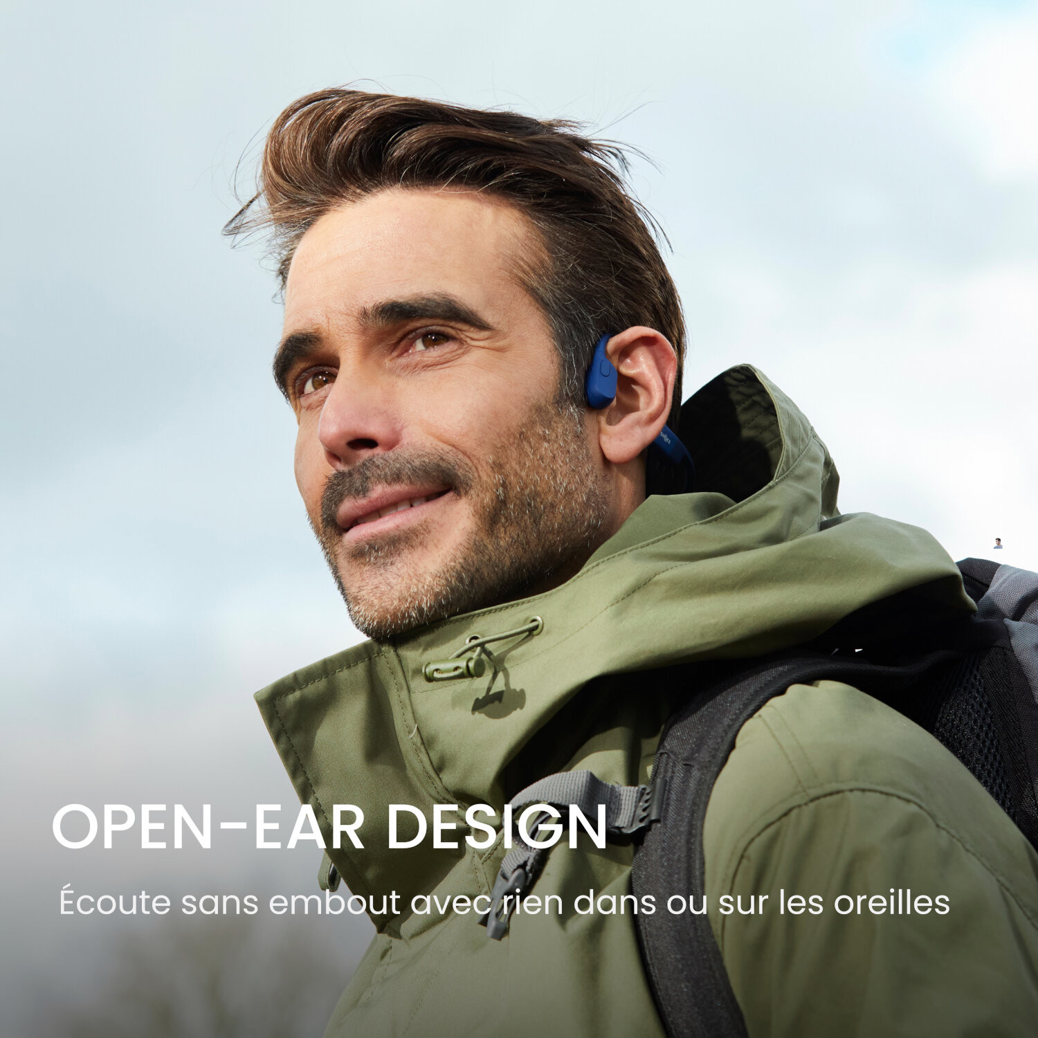 Homme portant des écouteurs à oreilles ouvertes. Il porte une veste verte et sourit. Extérieur.