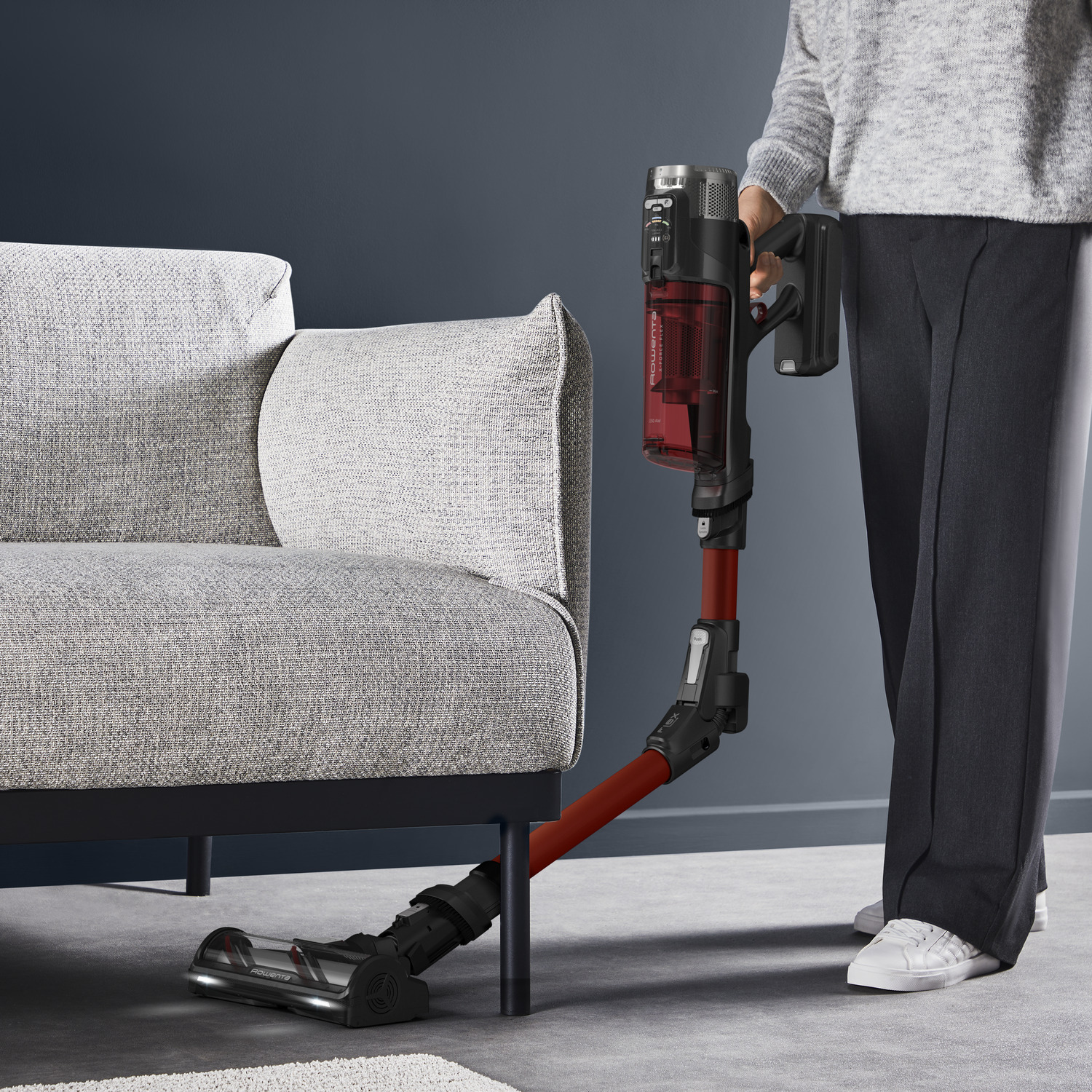 A person using a red vacuum cleaner near a gray sofa.