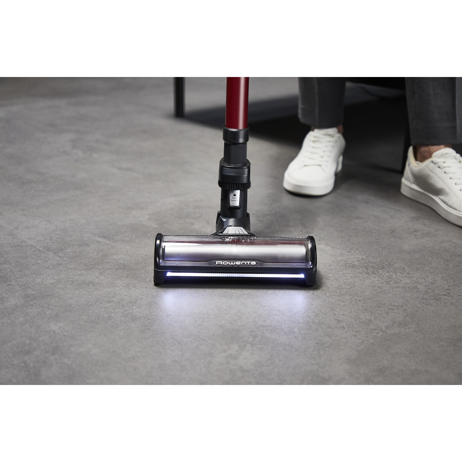 Vacuum cleaner head on the floor. It has a red handle and white shoes visible in the background.
