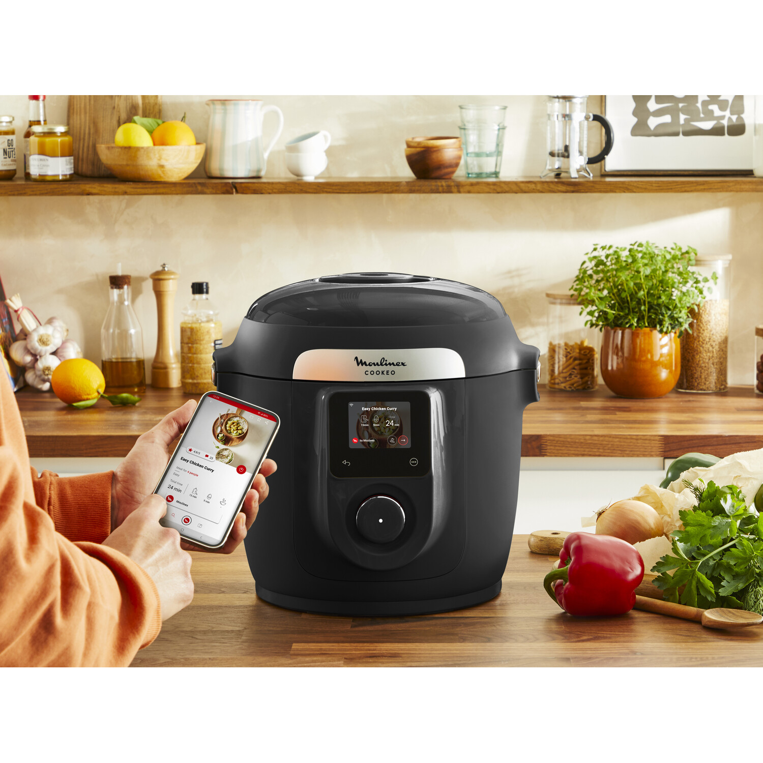 Personne utilisant un smartphone pour contrôler un multicuiseur intelligent Moulinex Cookeo noir sur bois.