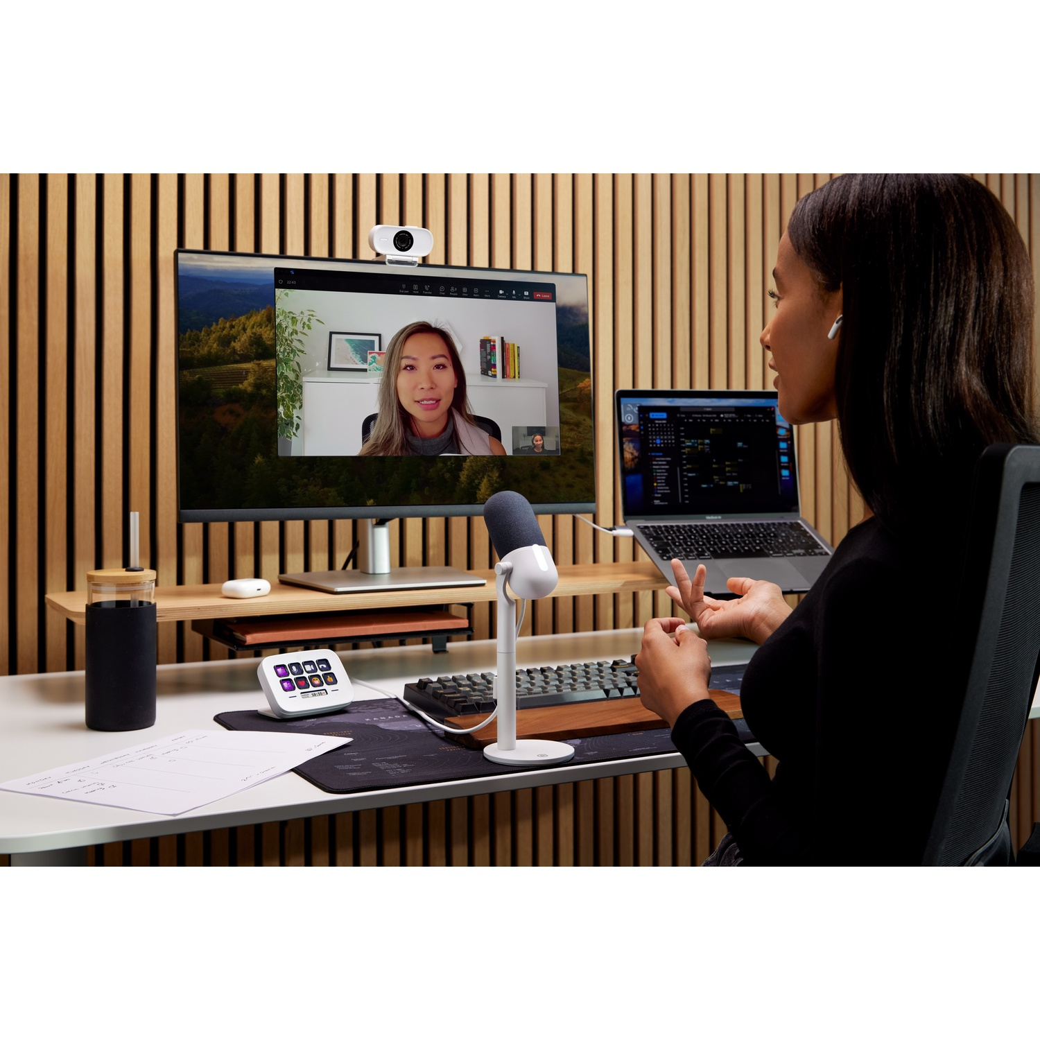 A woman on a video call on a monitor. A laptop, mic, and desk accessories are visible.