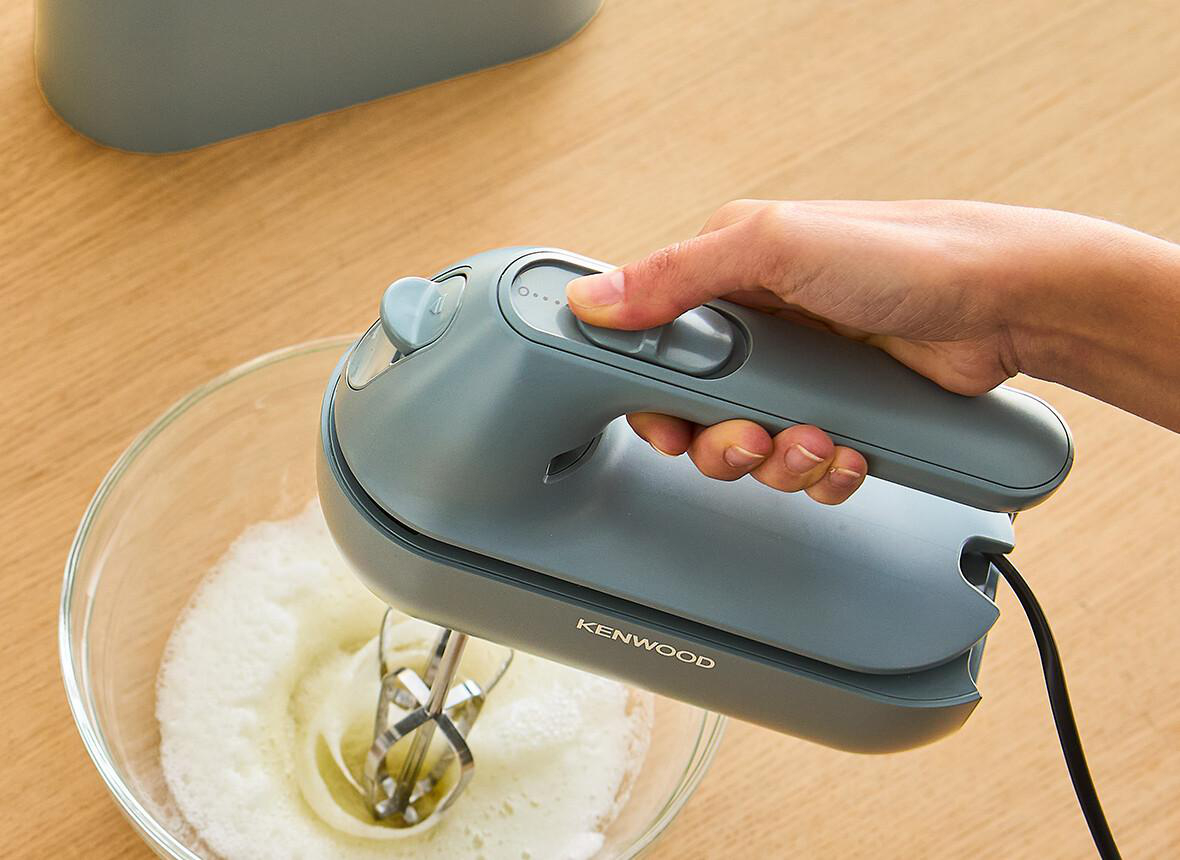 Eine Person benutzt einen grauen Kenwood Handmixer in einer Glasschüssel.