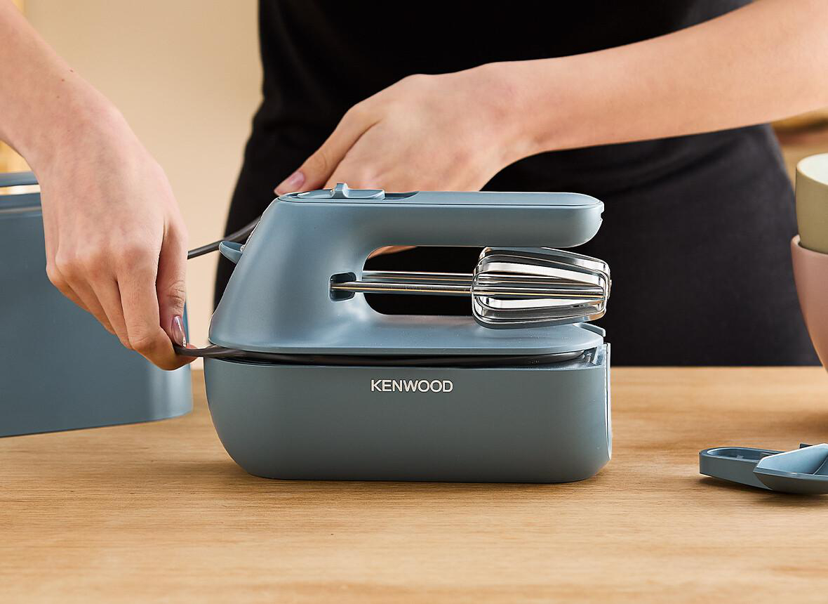 Person hält einen grauen Handmixer mit Drahtquirl auf einem Holztisch in der Nähe eines blauen Behälters.