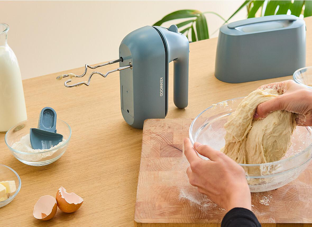 Person knetet Teig in einer Glasschüssel, mit Mixer und Zutaten auf einem Holztisch.
