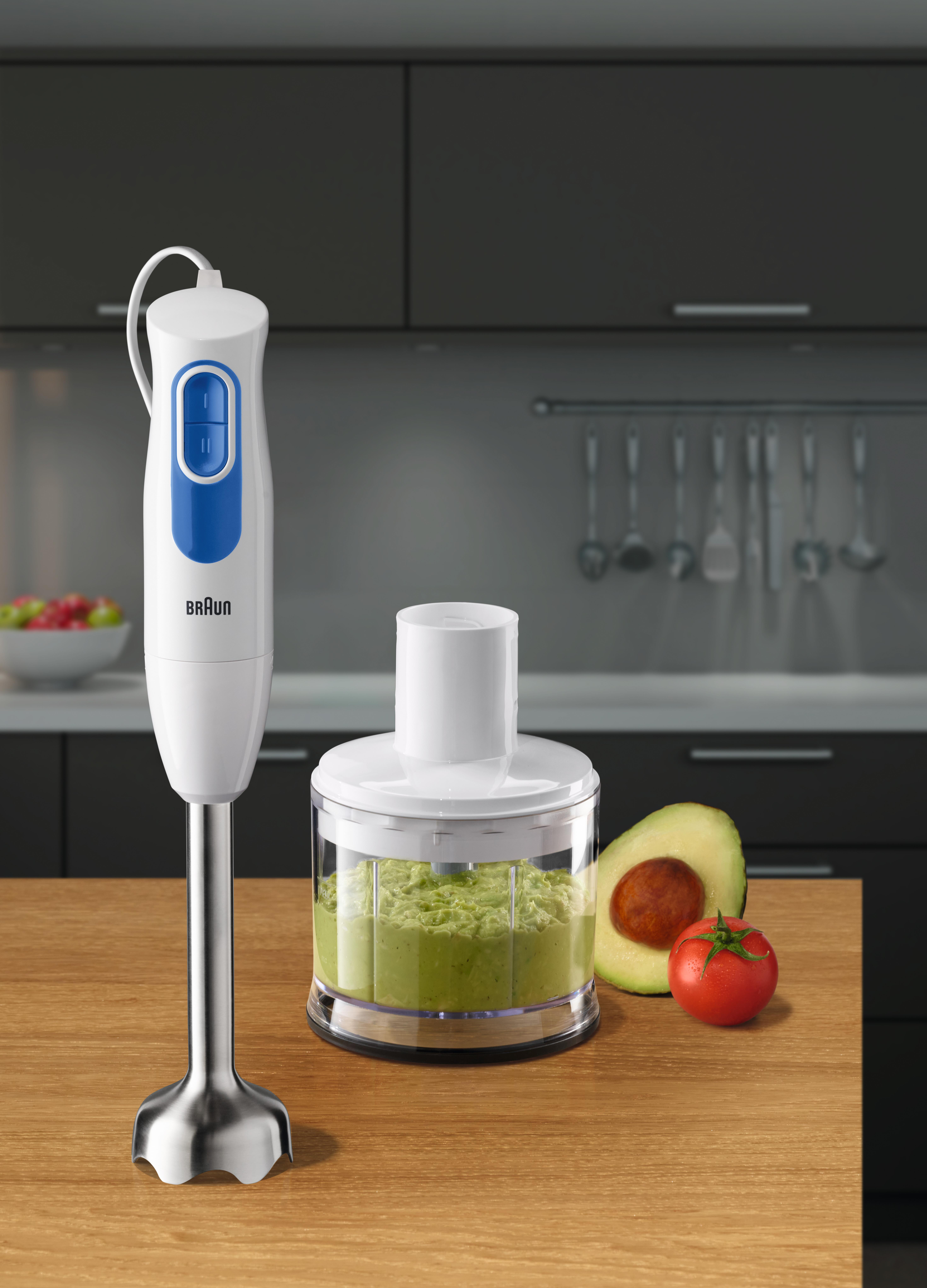 Weißer und blauer Braun-Handmixer, Behälter mit grüner Mischung, Avocado-Hälfte und Tomate.